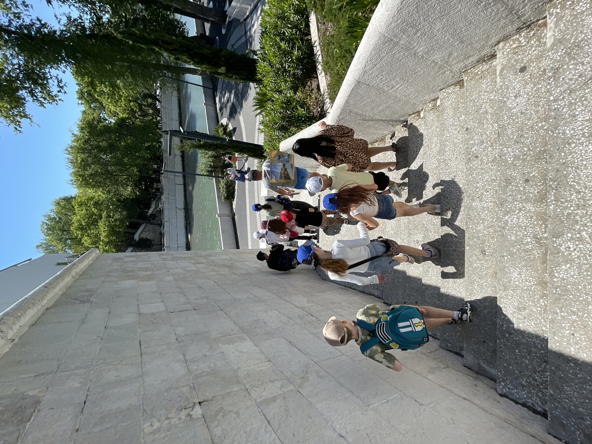 A group of approximately ten to twelve individuals, appearing to be mostly children and young adults, are captured walking upwards on a wide, sunlit stone staircase. The person closest to the foreground is a child wearing a camouflage-patterned long-sleeved shirt, a beige baseball cap, and a blue backpack adorned with white stripes and the number "1". Further up the stairs, other group members are dressed in casual clothing; notable among them is a person in a leopard print dress with dark hair, and several others wearing baseball caps of various colors. One individual appears to be carrying a framed object, possibly a picture or small canvas.
To the left of the staircase, a stone wall borders a calm body of water, resembling a canal or river, reflecting the bright blue sky. Beyond the waterway, a lush landscape of green trees and dense foliage lines the banks, with glimpses of distant buildings. The strong, elongated shadows cast by the people indicate bright, clear weather and a high sun, suggesting it is midday or early afternoon. The scene depicts a casual activity, likely a tour or outing, in an outdoor, picturesque setting. The visible elements, including the stone architecture and a waterway amidst greenery, align with a scenic urban or park environment, consistent with the specified location of Sochi, Russia.
