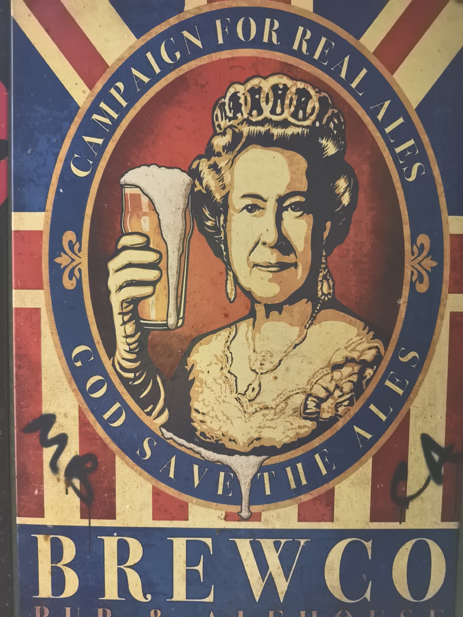 The image depicts a pub sign for "BREWCO," a pub and alehouse. The sign features a stylized portrait of Queen Elizabeth II holding a pint of beer. The background of the portrait is a red circle, which is superimposed onto a Union Jack design. Around the circle, text reads "CAMPAIGN FOR REAL ALES" at the top and "GOD SAVE THE ALES" at the bottom. Some graffiti is visible near the bottom corners. The location in the city of Noviy, Russia is not determinable from the image.