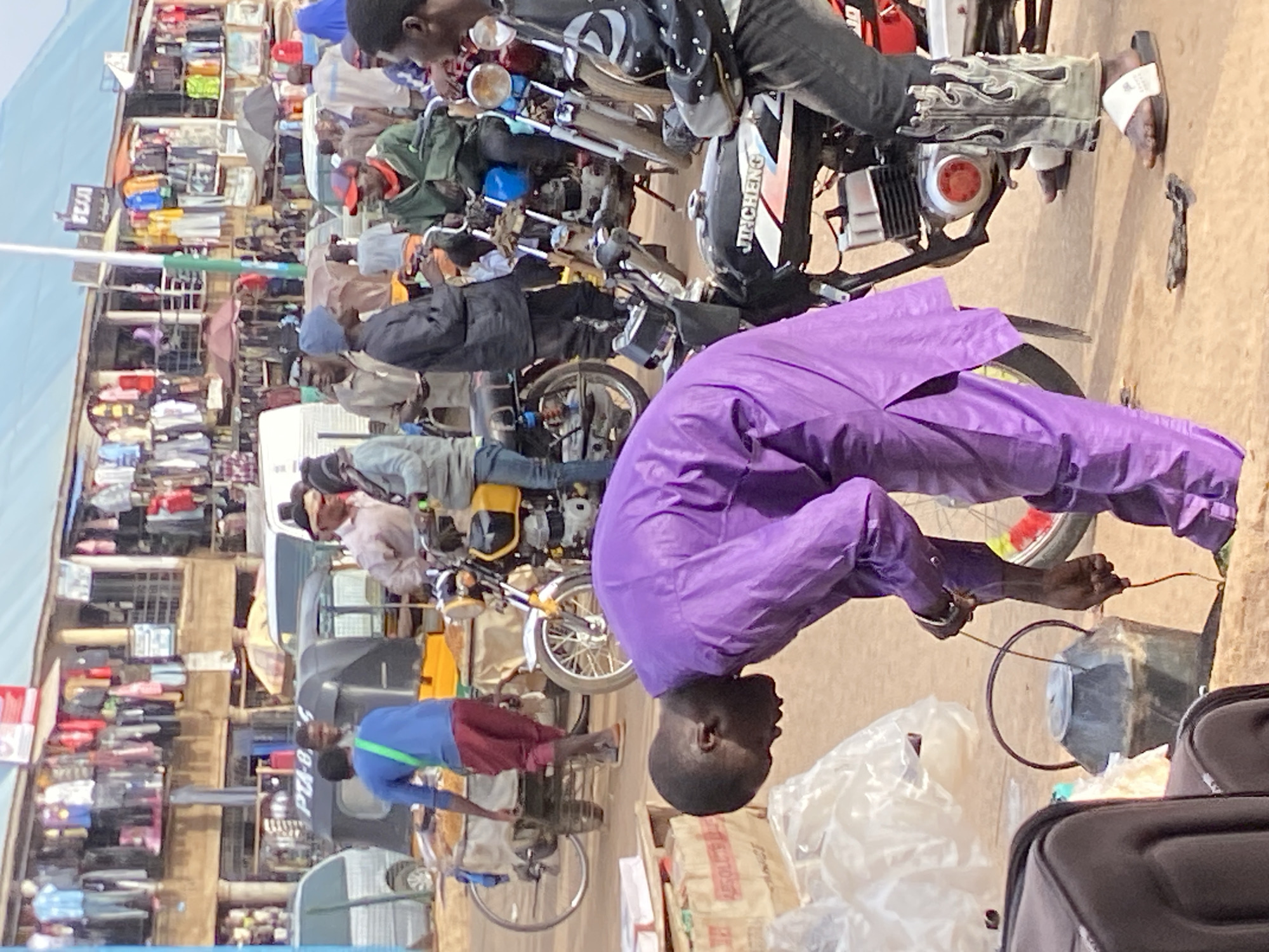 This is a medium shot photograph taken outdoors in Bukuru, Nigeria. The scene depicts a bustling street market with several people and motorcycles. The foreground is dominated by a man wearing a vibrant purple robe, bent over and appearing to interact with something on the ground. Next to him, a large black container with a handle and lid sits on the ground, and several bags, possibly luggage, are visible. A motorcycle with the brand name "JINCHENG" is parked nearby.
In the mid-ground, other individuals are seen standing or sitting on motorcycles. One man in a blue shirt and red trousers stands near a tricycle, which is laden with goods. Another motorcycle with a yellow tank is parked to the left.
The background reveals a row of shops with clothing and other items displayed outside. The architecture suggests a market setting. The overall impression is one of a lively commercial area in Bukuru. The lighting suggests it is daytime, with sunlight casting shadows. The ground is unpaved, indicating a typical street environment.