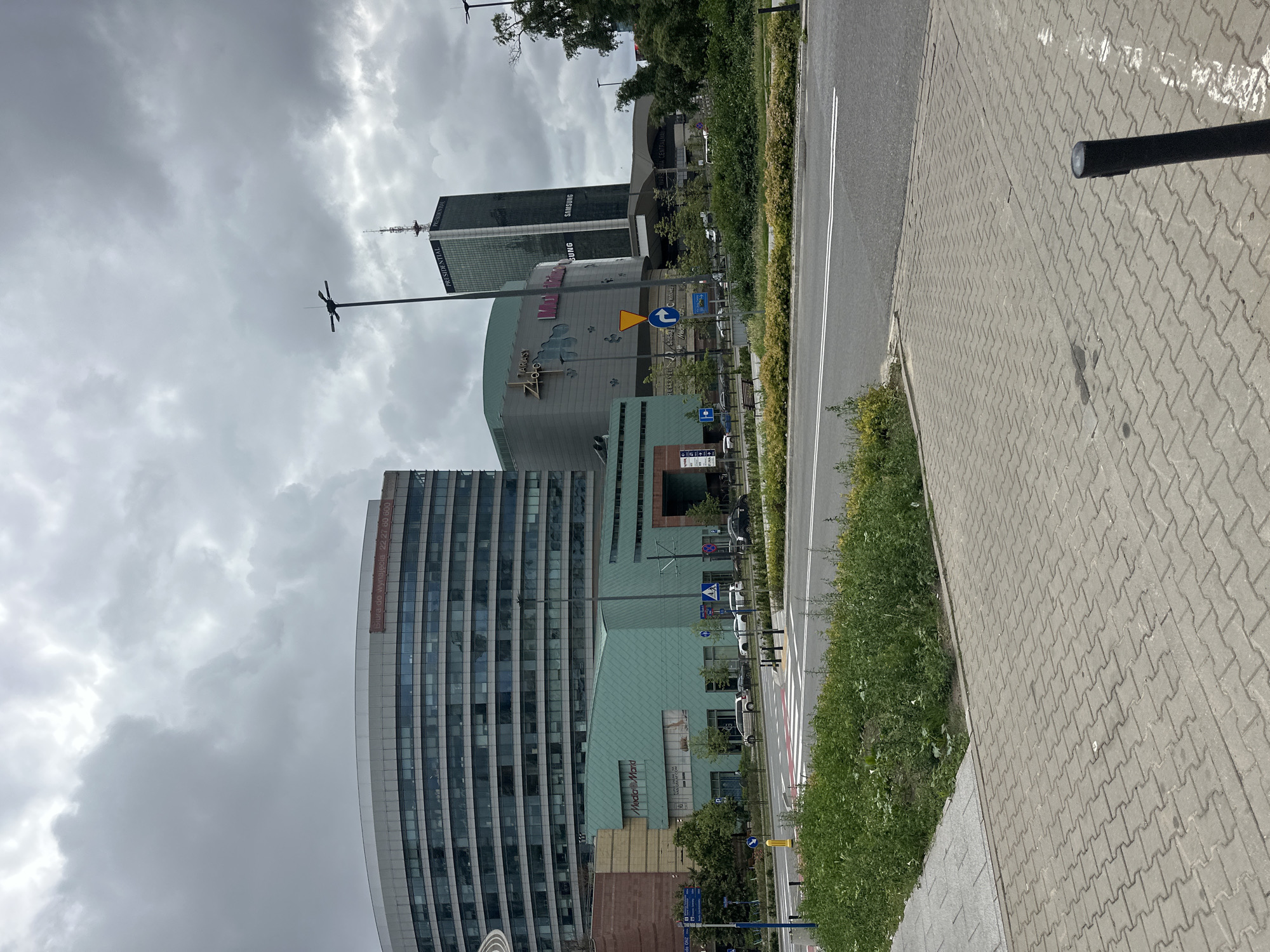 The image depicts an exterior view of a city street in Warsaw, Poland, on a cloudy day. In the foreground, a paved sidewalk made of interlocking gray pavers leads to a street with visible lane markings and a crosswalk. To the left of the sidewalk, a patch of green grass and weeds is visible.
Dominating the background are several large, modern buildings. On the left, a curved, multi-story building with a glass facade is partially visible, with a banner displaying "Biura do wynajęcia 22 27 80 800" (Offices for rent). Adjacent to it, a shorter, light green building with a sloped roof features a prominent "Media Markt" sign. Further to the right, a tall, dark glass skyscraper stands, with "PRESIDENTIAL" and "SAMSUNG" signage visible. Another building with a curved roof and signs that appear to read "TARASY ZŁOTE TARASY" and "Multikino" is also present. The sky is heavily overcast with gray clouds. Streetlights, traffic signs, and parked cars are also part of the urban landscape. The scene appears to be a commercial or business district.