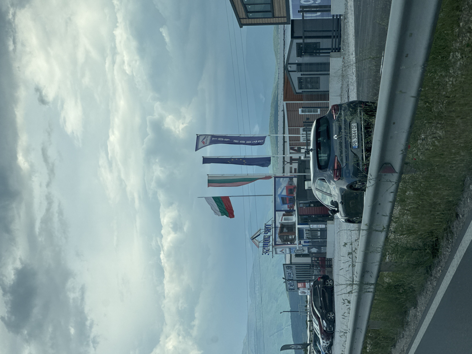 The image shows a scene from near Plovdiv, Bulgaria. The photo, likely taken from a moving vehicle, captures a cloudy sky above a commercial area alongside a road. Several flags are visible, including the Bulgarian flag, the European Union flag, and a flag advertising "Top House". Prefabricated buildings or modular homes are displayed in the area. Cars are parked along the roadside. A guardrail is on the right. The car's license plate number is PB 3044 CE.