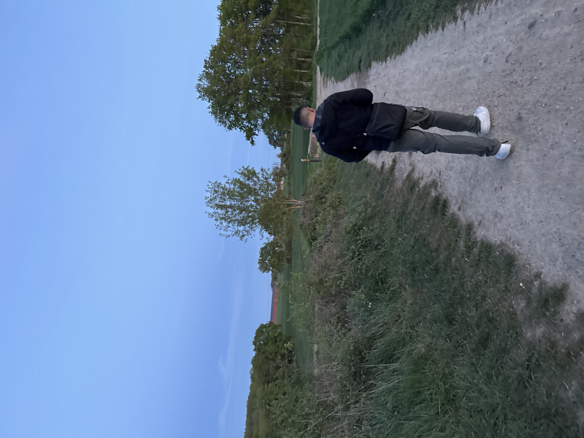 This image, taken in Gometz-le-Châtel, France, captures an outdoor scene at what appears to be late afternoon or early evening. The photograph is oriented vertically, with the natural horizon running down the left side of the frame.
The main subject is a person, seen from behind, standing on a light-colored dirt path that stretches diagonally from the bottom right towards the top left. The individual is a male, wearing a dark (likely black) jacket, dark green or khaki cargo pants, and white athletic shoes. A black shoulder bag is visible across his left shoulder. He has short, dark hair.
The path is bordered by green grass and various plants, with darker, more overgrown bushes and foliage on the immediate right. To the left of the path, there's a wider area of green grass and several trees of varying sizes and foliage densities. In the further background, a field of green is visible, and faint reddish structures, possibly roofs or buildings, can be discerned on the distant horizon line. The sky, occupying a significant portion of the left side of the image, is a clear, serene light blue, indicative of a peaceful time of day with soft, diffused lighting. There are no visible clouds. No text is present in the image.