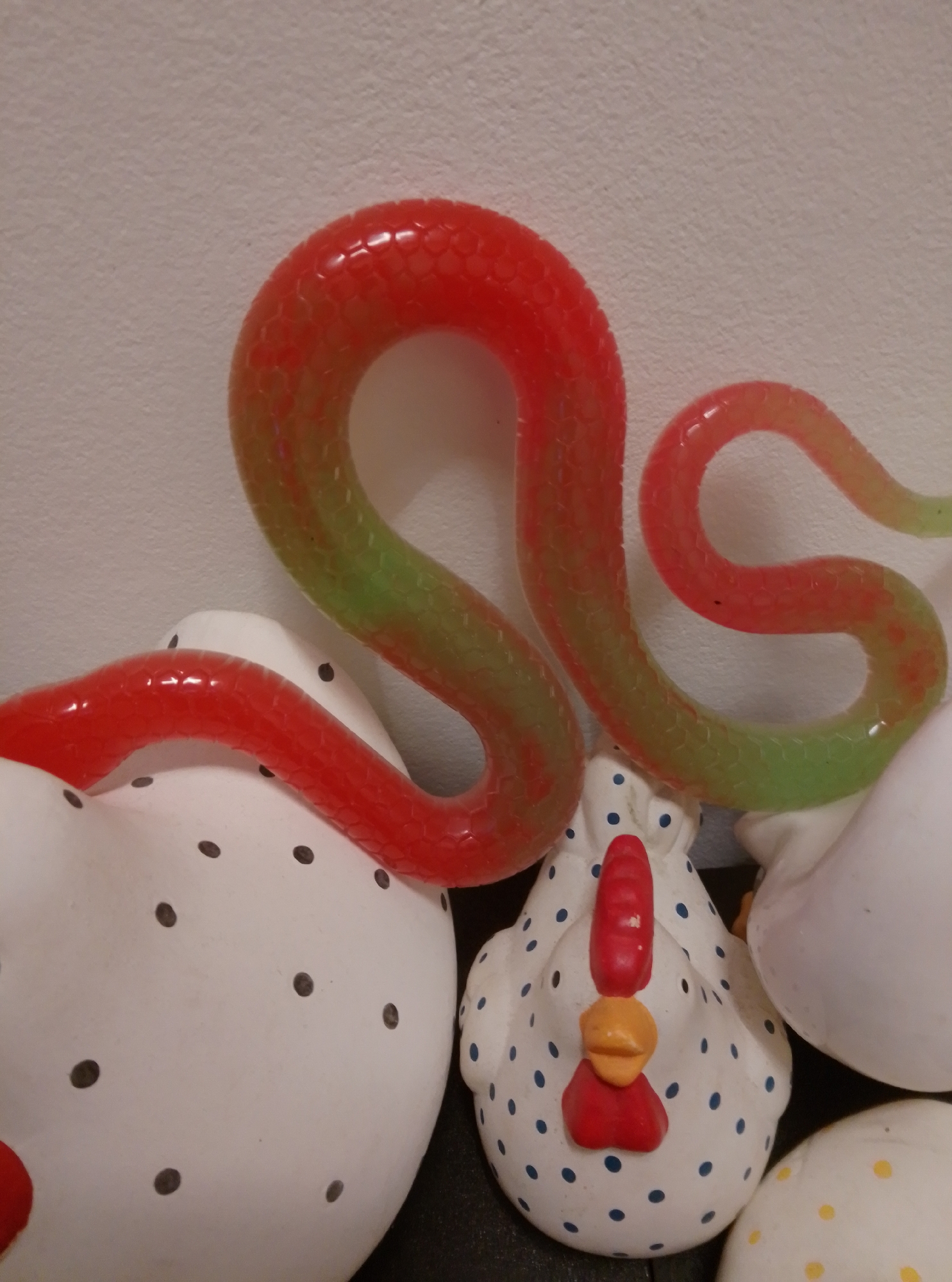 A plastic toy snake, colored red and green, is draped over three ceramic chicken figurines and an egg. The snake is coiled and appears to be positioned on top of the chickens and egg. The chickens are white with dark polka dots. One chicken has a red comb and wattle, and an orange beak. Another chicken, to the right, is mostly obscured. The egg, in the lower right corner, is white with yellow polka dots. The background is a plain, textured white wall. The scene is indoors, with artificial lighting. The image does not provide any information that would allow for its location to be determined, including in the city of Wedel, Germany.