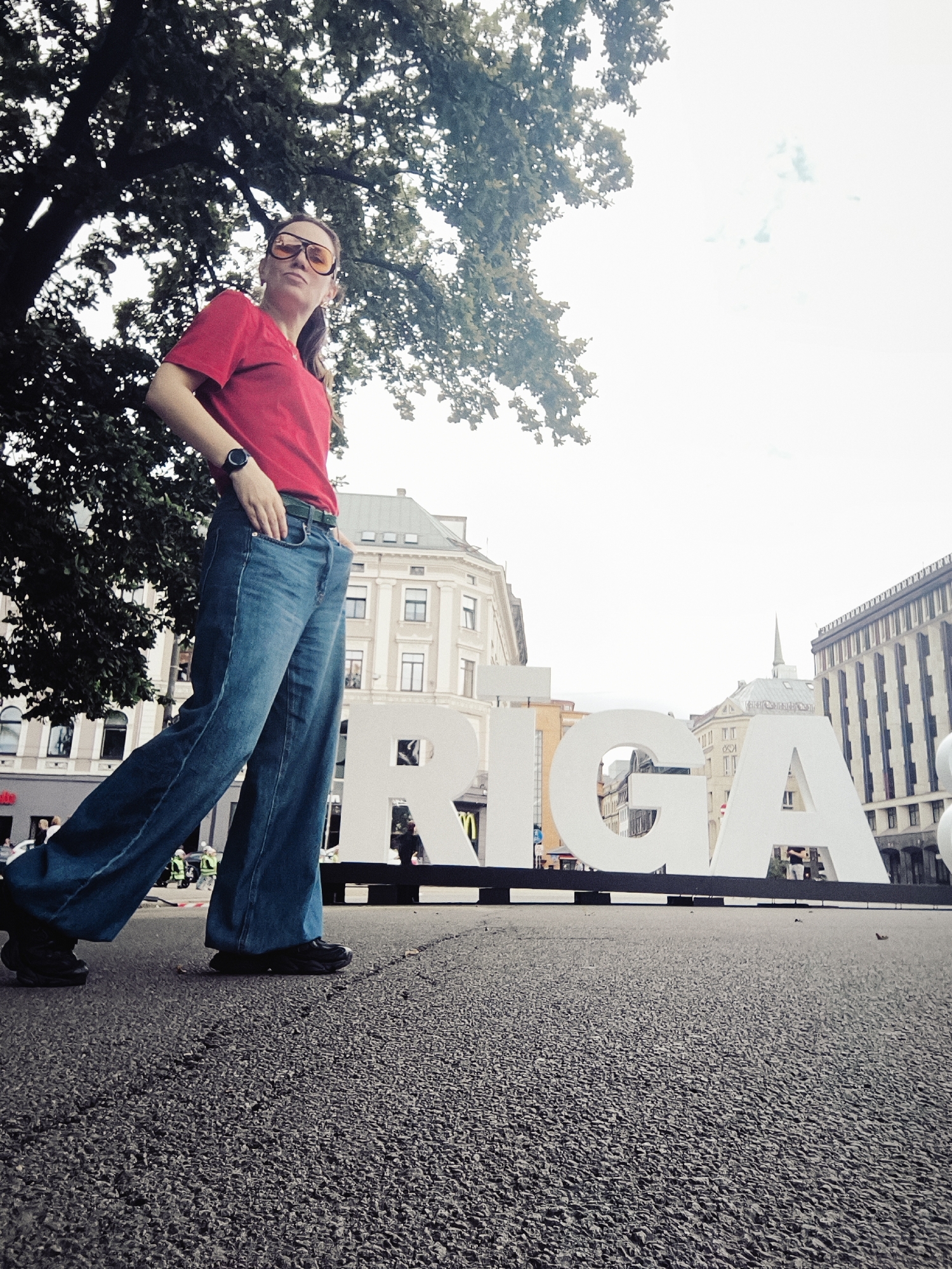 A low-angle shot shows a woman walking past a large, white "RIGA" sign in a city square. The woman, seen from the waist down, wears a red t-shirt, blue bell-bottom jeans, and black sneakers. She has her left hand in her pocket. The "RIGA" sign is monumental, with each letter standing several feet tall. Behind the sign, a building with classical architecture is visible. The street is paved with dark asphalt. The sky is overcast, and a large tree with green leaves partially shades the scene.