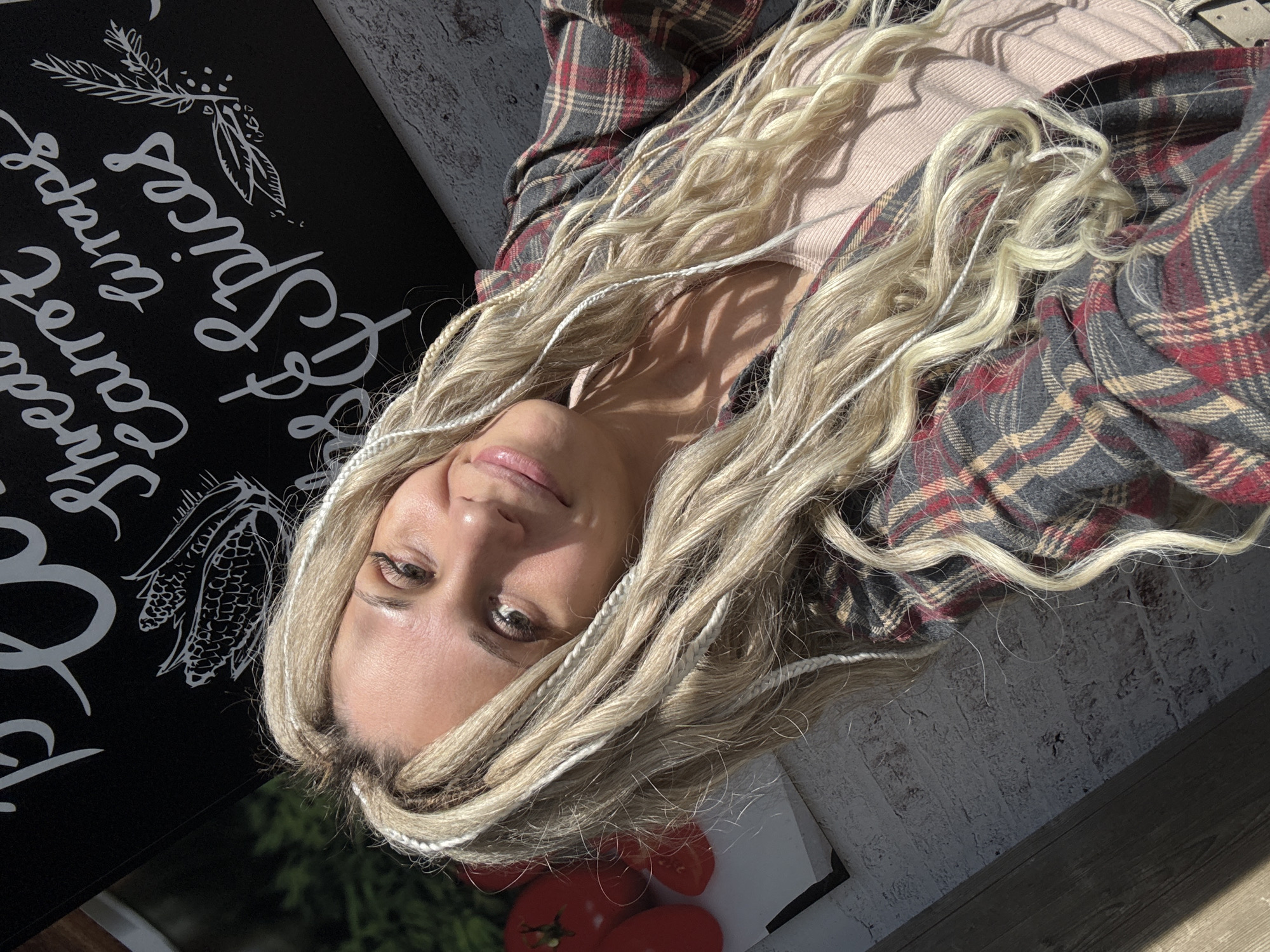 A woman with blonde hair and white braids is centered in the frame. She is wearing a tan shirt under a red and gray plaid flannel. To the left of the woman is a black sign with white cursive lettering that says "Sweet Carrot Curry & Spices". Underneath the sign and to the right of the woman's face, there are several red tomatoes. The background appears to be an outdoor setting near a white brick wall and wooden flooring. Based on the sign and the visible location information from the user prompt, the location is likely within Rusanovo, Russia.