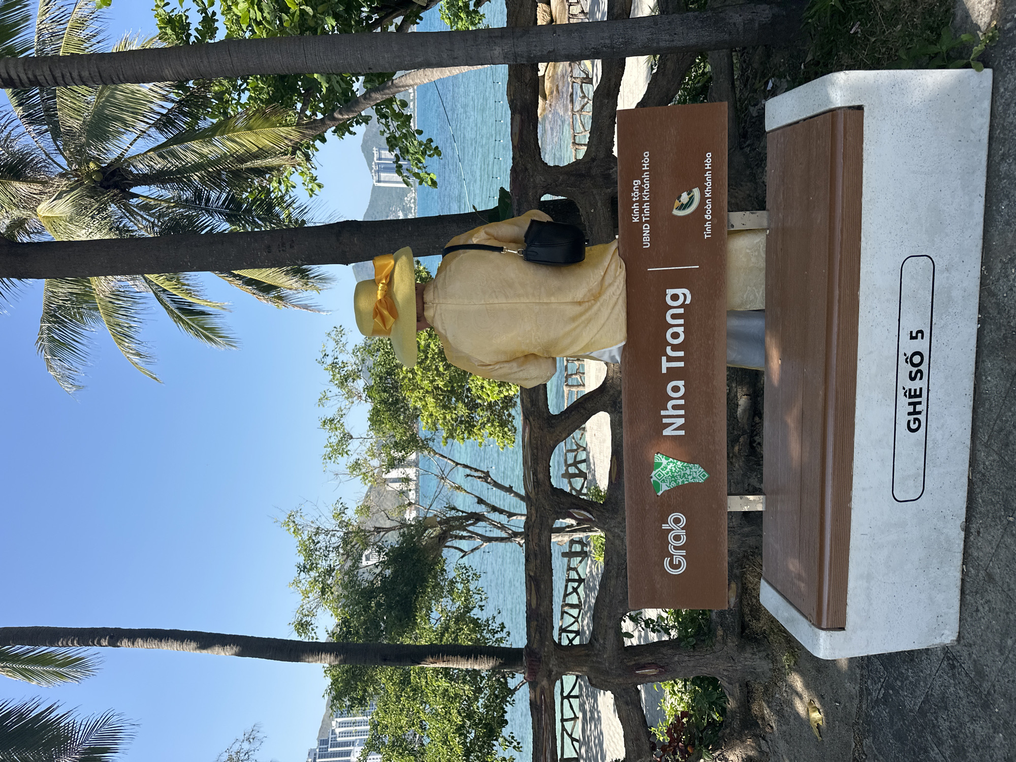 The image presents an outdoor coastal scene on a clear, sunny day in Nha Trang, Vietnam. A single individual is seated on a public bench, observed from behind, facing a calm, blue-green sea. The person wears a light-yellow, long-sleeved garment, possibly an Ao Dai top, and a wide-brimmed yellow straw hat with a large yellow bow. A black shoulder bag with a strap hangs from their right shoulder.
The bench has a concrete base and a wooden-slat seat and backrest. A brown plaque on the backrest displays "Grab Nha Trang" with a green map-like emblem, and below it, "Kính tặng UBND Tỉnh Khánh Hòa" and "Tỉnh đoàn Khánh Hòa" in white text. The front of the concrete base is marked with "GHẾ SỐ 5".
The immediate foreground includes the bench and the ground, which appears to be paved. Beyond the bench, a decorative, dark-colored barrier with organic, branch-like patterns separates the area from a walkway closer to the water. Numerous green trees and bushes are visible, along with several tall palm trees that frame the scene. In the midground, beyond the barrier, string lights are visible stretched above the water.
The background features distant mountains or hills with multiple modern high-rise buildings along a far coastline. The sky above is clear and bright blue.