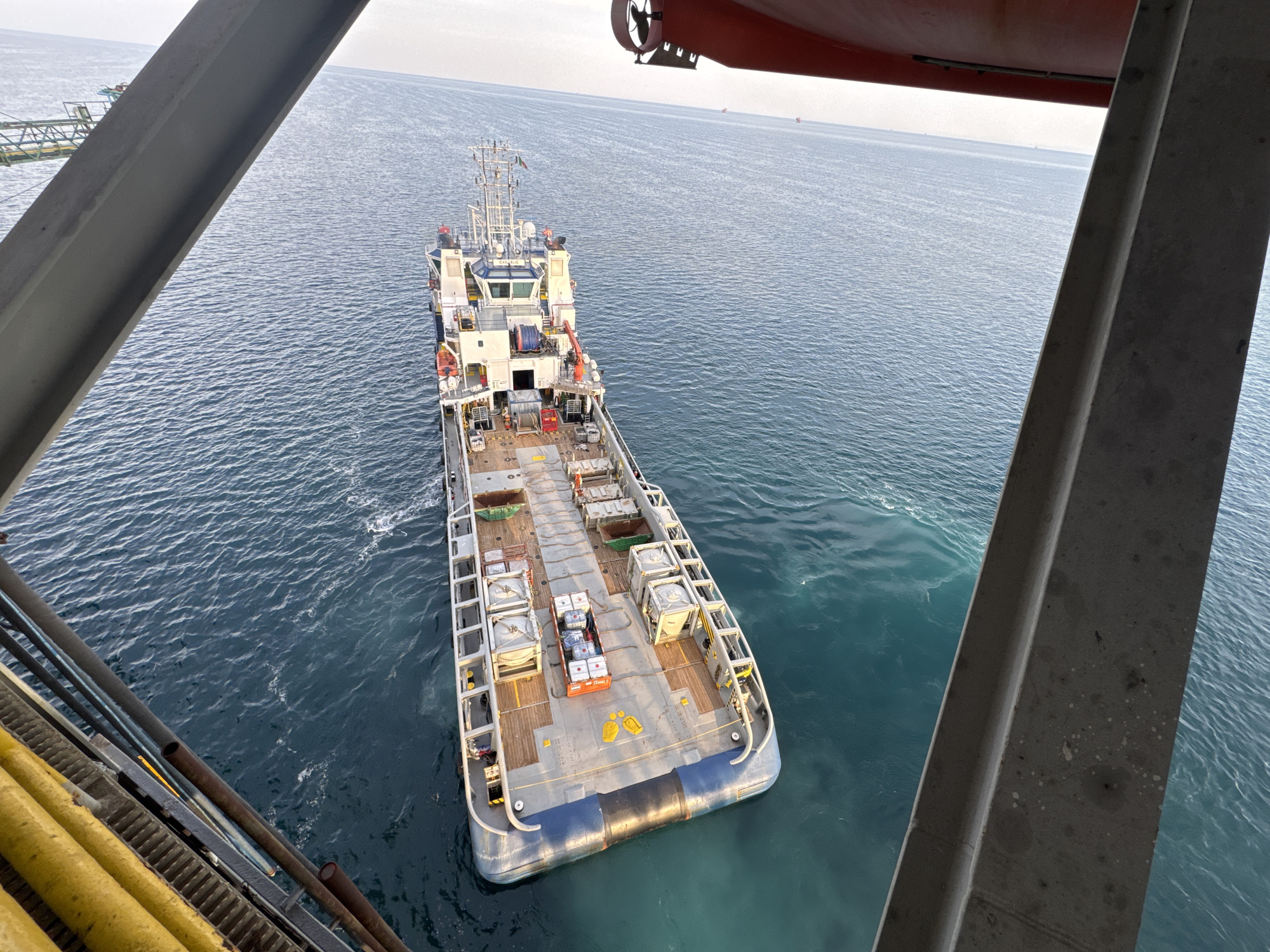 This is a high-angle, eye-level, medium shot captured on a boat, likely a supply vessel or a large barge, sailing on a calm, blue ocean. The vessel is positioned in the center of the frame, viewed from the stern towards the bow. Its deck is laden with various equipment, including several large, rectangular containers, some of which appear to be covered. In the middle of the deck, a smaller orange container holds several white boxes. The vessel has a prominent bridge structure towards the bow and appears to be equipped with cranes and winches.
In the foreground, a portion of another vessel or platform is visible, with thick yellow pipes and metal structures occupying the left side of the image. The water is a deep blue, with slight ripples indicating calm conditions. In the distance, the horizon is visible, and several other vessels, appearing as small specks, can be seen scattered across the sea. The sky is overcast, suggesting a mild or cloudy day. The overall scene depicts a working environment at sea, likely during the daytime. There is no discernible text visible in the image.