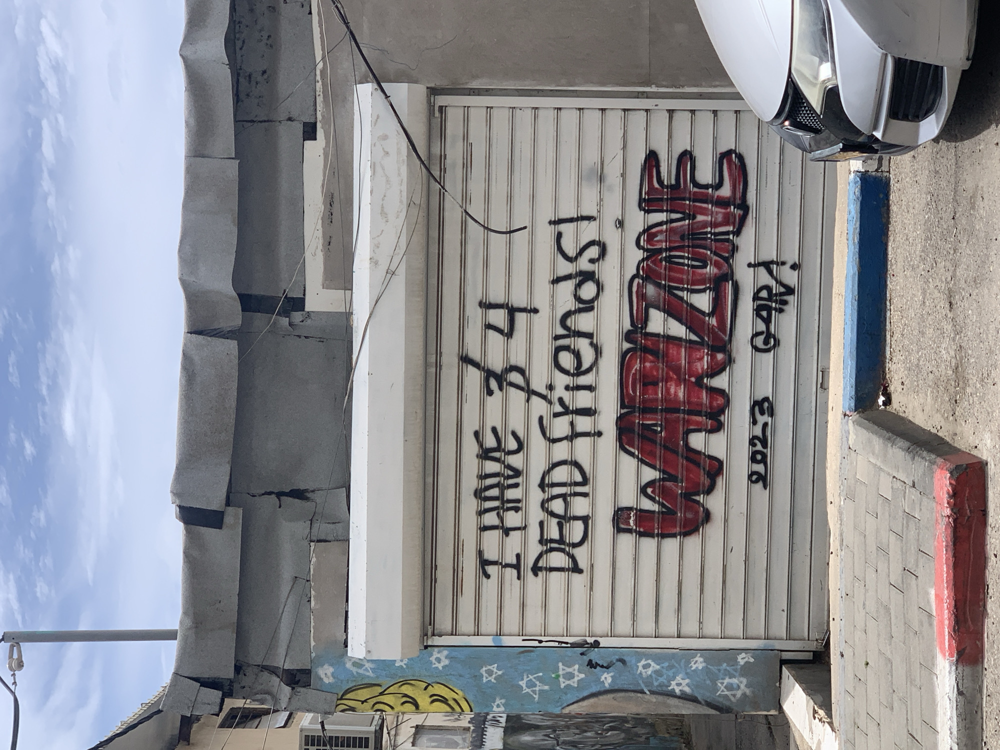 A closed, horizontally ribbed metal security gate is covered in graffiti. The graffiti reads "I HAVE 3/4 DEAD FRIENDS!" in black block letters above the red and black graffiti tag "WARZONE." Underneath, also in black, is the year "2023" and the name "GARA." The gate is white and has a few dangling wires to its left. The gate appears to be part of a building along a street. The car is also partially visible in the top right corner of the shot. The image was taken outdoors, in Tel-Aviv.