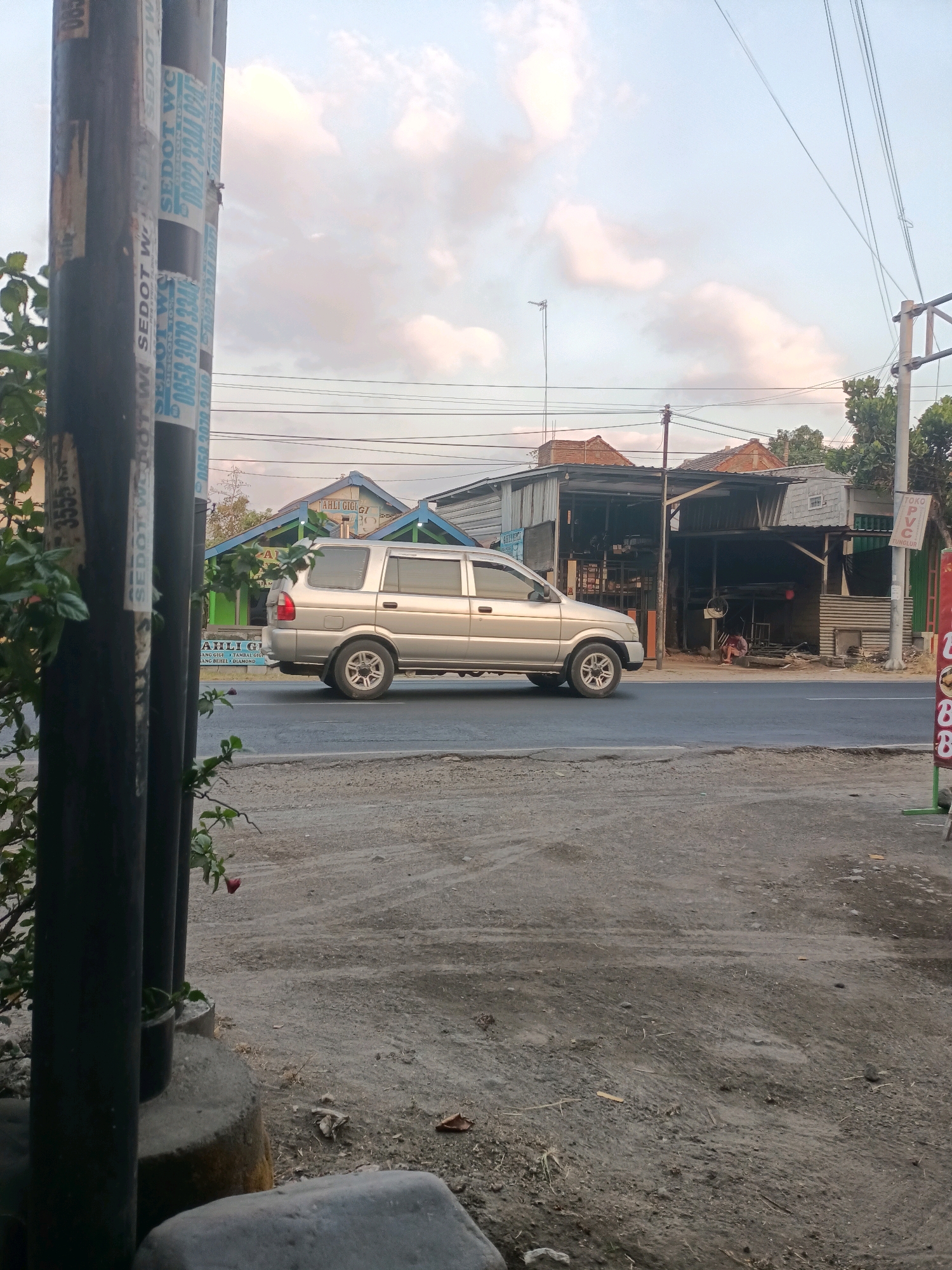 The image captures a street scene in Badas, Indonesia, under a cloudy sky. A silver SUV is parked on the right side of the road, facing left. To the right of the SUV, a building with a corrugated metal roof and open sides appears to be a shop or workshop. A person is seated in front of this building. On the left side of the image, a large black pole with several stickers is visible, partially obscuring a leafy plant. In the background, residential buildings with tiled roofs can be seen. The road surface is asphalt, and the foreground consists of a dirt and gravel area. A red flower is blooming on a vine attached to the black pole.