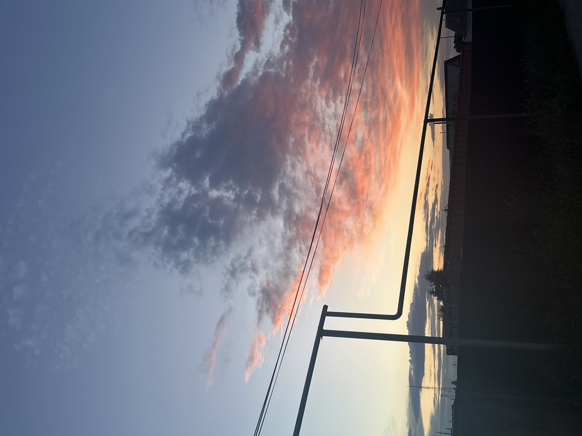 This is a photograph of a sunset in Tobyl, Kazakhstan, taken from a roadside perspective. The sky is a dominant feature, with a clear gradient of colors from a pale blue at the top to a soft yellow and orange near the horizon, indicative of twilight. Clouds are scattered across the sky, some a deep grey and others illuminated by the setting sun with vibrant pink and orange hues.
In the foreground and mid-ground, utility poles and power lines are visible, silhouetted against the colorful sky. These objects suggest an urban or semi-urban setting. To the right of the frame, a dark silhouette of vegetation and possibly a fence line runs along the edge of the road, further reinforcing the roadside context. In the distance, faint outlines of buildings and structures can be seen, also silhouetted against the sky.
The overall mood of the image is serene and peaceful, evoked by the soft colors of the sunset and the quiet, empty scene. There is no text visible in the image. The time of day is clearly sunset or twilight. The weather appears clear, with the clouds adding visual interest to the sky.
