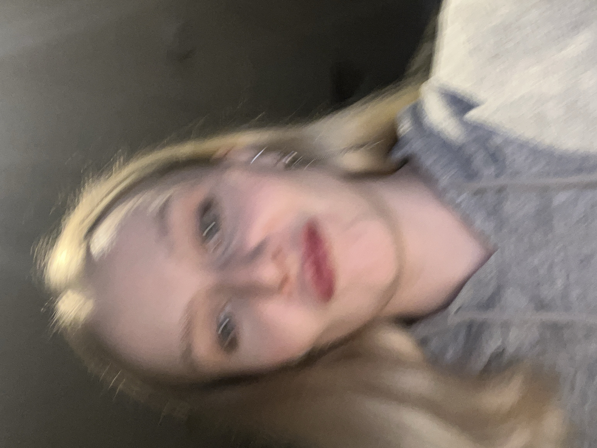 A close-up, slightly blurred, selfie-style photograph depicts a young woman with fair skin and blonde hair. She is wearing lipstick. She appears to be wearing a gray hooded sweatshirt. The background is dark. The location in Khimki, Russia is not discernible from the image.