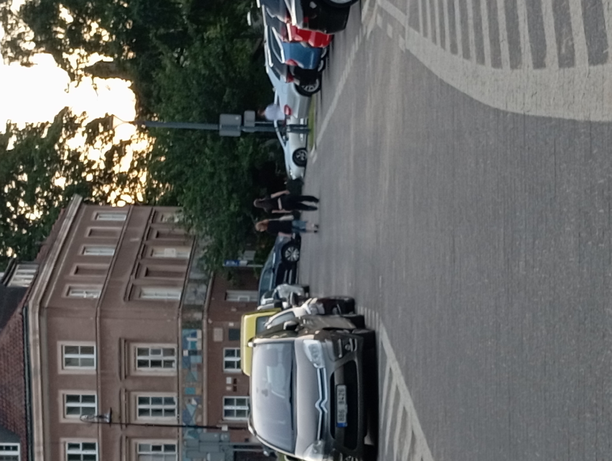 In Warsaw, Poland, an outdoor scene captures a street view with a multi-story building on the left and parked cars lining the right side of a paved area. The building, constructed with a reddish-brown facade and multiple windows, exhibits architectural details consistent with older European city structures. In the foreground, a dark grey car, identified by its license plate "886 8426", is parked on the right. To its left, a yellow vehicle is partially visible. Further down the street, a line of cars, including a blue and a red sedan, are parked. Two women, one in black attire and the other in a dark top and lighter pants, are walking away from the viewer towards the right side of the frame. A tall lamppost stands near the building on the left. The setting appears to be an urban environment, possibly a parking lot or a street in a city. The lighting suggests it is late afternoon or early evening.