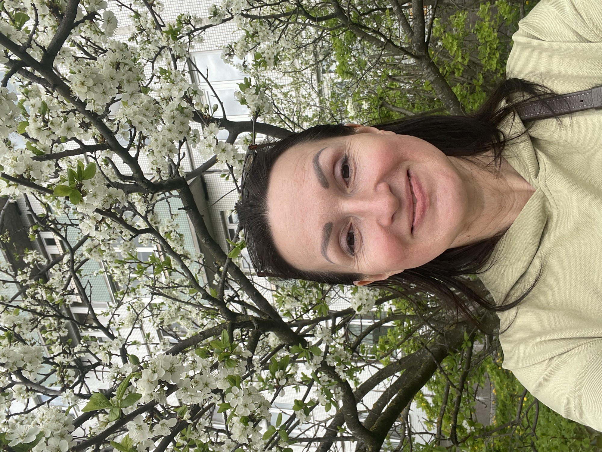 A woman is posing for a selfie outdoors. She has brown hair, fair skin, and is wearing a light green hoodie. She's positioned in front of a tree in full bloom with white flowers. Behind the tree, a light-colored building with multiple windows is visible. The location is Saint Petersburg, Russia.