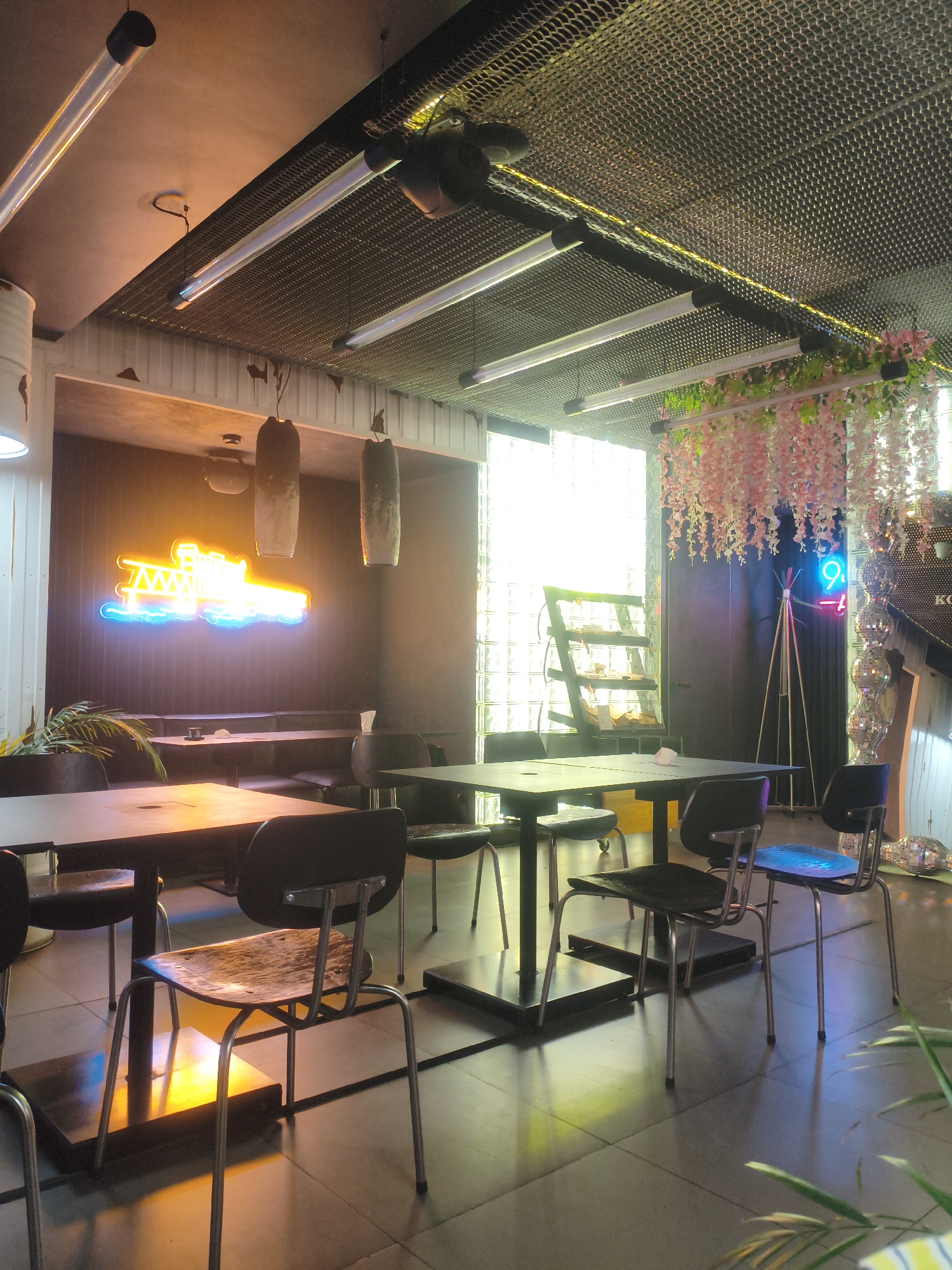 The image depicts the interior of a cafe or restaurant, possibly located in Kremenchuk, Ukraine, based on the user's prompt. The space features a modern, industrial aesthetic with a suspended metal mesh ceiling and exposed lighting fixtures. Tables and chairs are arranged throughout the room. A prominent neon sign shaped like a construction crane, in yellow and blue, is mounted on a dark wall. Decorative elements include hanging artificial pink flowers and a large, multi-faceted disco ball structure. Natural light streams in from a large window composed of glass blocks. The overall atmosphere appears to be dimly lit, with warm ambient lighting from the neon sign and overhead fixtures, casting reflections on the tiled floor. There are no people visible in the scene.