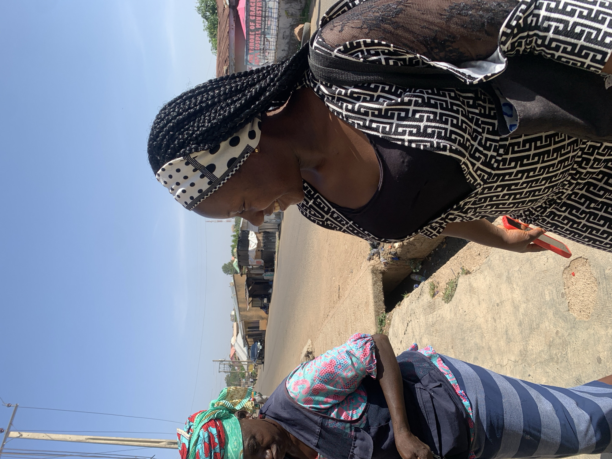 The image shows two Black women standing on a concrete sidewalk in Jos, Nigeria. One woman is wearing a black-and-white patterned shirt and a polka-dotted headscarf, and she is looking down with a smile. The other woman is wearing a green and turquoise patterned head wrap and a blue-and-pink patterned shirt under a navy vest and a striped dress. A street and some buildings are visible in the background. The sky is clear and blue.