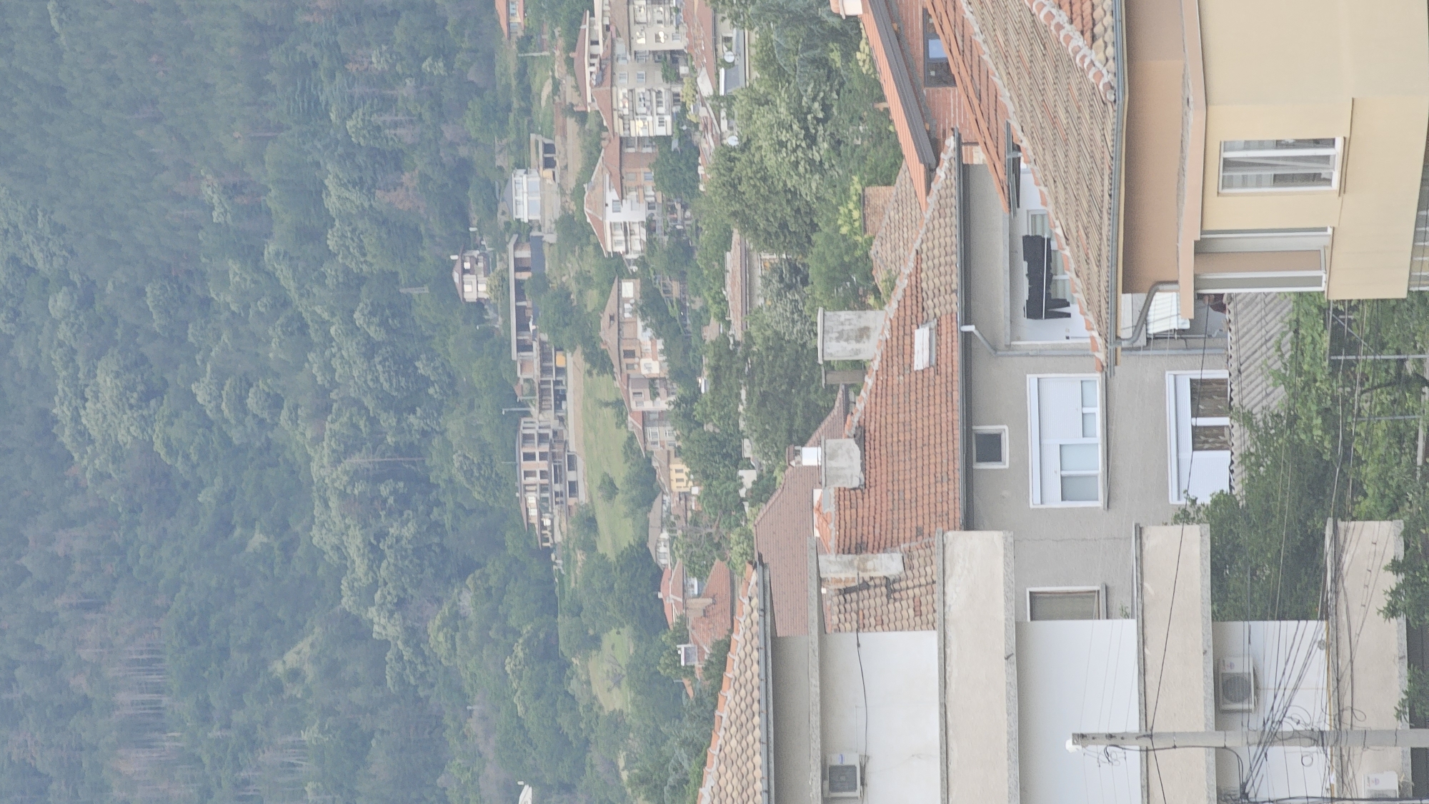 The image depicts a view of a residential area in Karlovo, Bulgaria, nestled against a hillside covered in dense green foliage. The foreground features the rooftops of several houses, characterized by terracotta tiles and brick chimneys. One building in the lower left exhibits a white facade with an air conditioning unit. Further into the scene, more houses are visible, varying in architectural style and color. Some are older, with visible brickwork, while others are painted in lighter hues like white and beige. Multiple windows, some with white shutters, are scattered across the buildings. A few unfinished or under-construction buildings are present on the hillside, suggesting ongoing development. Power lines and poles are visible in the lower portion of the image, and trees and shrubs are interspersed among the houses, indicating a well-established residential neighborhood. The overall impression is a semi-urban environment with a strong natural backdrop of a wooded hill.