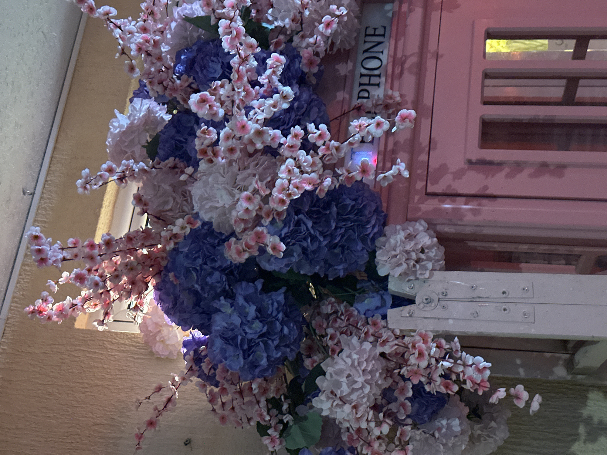 The image shows a close-up of an artificial floral arrangement in front of a pink structure, likely a decorative element resembling a phone booth. The flowers are a mix of large, dark blue and light pink hydrangeas and delicate pink cherry blossoms. The pink structure has the word "PHONE" visible on its upper section, suggesting it is indeed a stylized phone booth. The background is a textured beige wall. The lighting appears to be artificial, with a pinkish hue casting over the scene, particularly on the right side of the phone booth. There are no people or animals visible in the image. The location is identified as being in the city of Budva, Montenegro.
