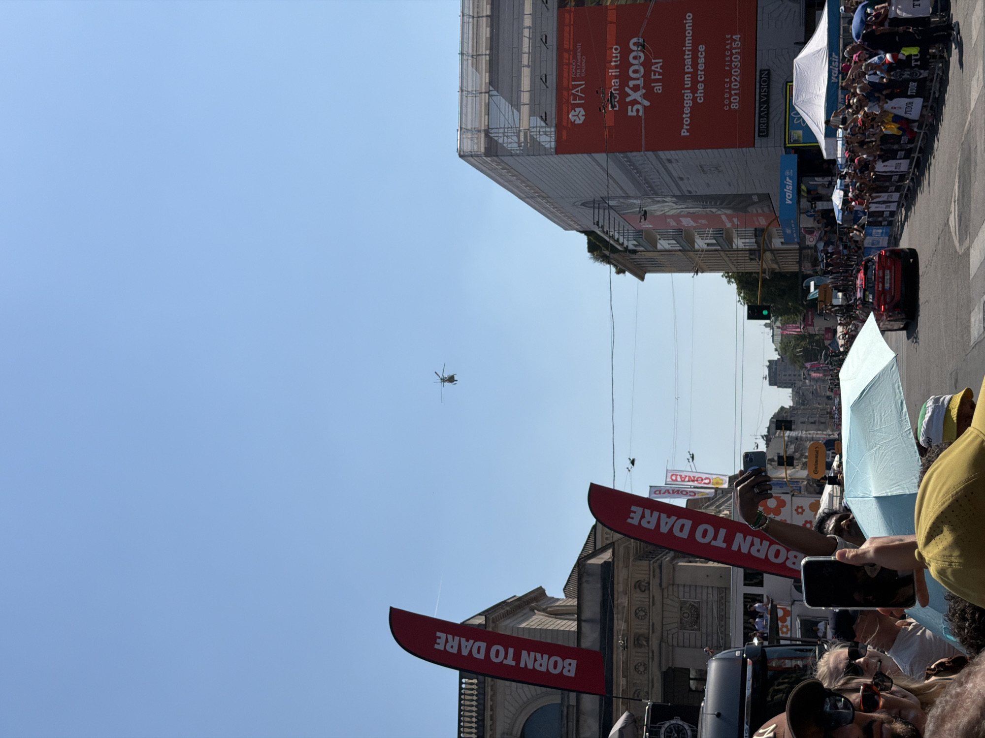 The image is a low-angle shot taken at ground level in what appears to be Milan, Italy. In the bright, clear sky is a helicopter, relatively small in the frame. The foreground is crowded with people, some holding up their phones to take photos or videos. Many people are partially obscured by umbrellas and signage. Several red banners emblazoned with the words "BORN TO DARE" are prominently displayed. Behind the crowd is a building, possibly a historical landmark given its architecture. On the right side of the image, a large advertisement covers the side of another building, featuring text in Italian regarding a "5x1000" initiative and promoting FAI - Fondo Ambiente Italiano. The presence of brands such as "valsir," "Continental," and "CONAD" suggest a commercial or sponsored event is taking place. The presence of barriers or fences indicates that the crowd is being managed. The scene overall suggests a public event or celebration occurring in an urban setting.
