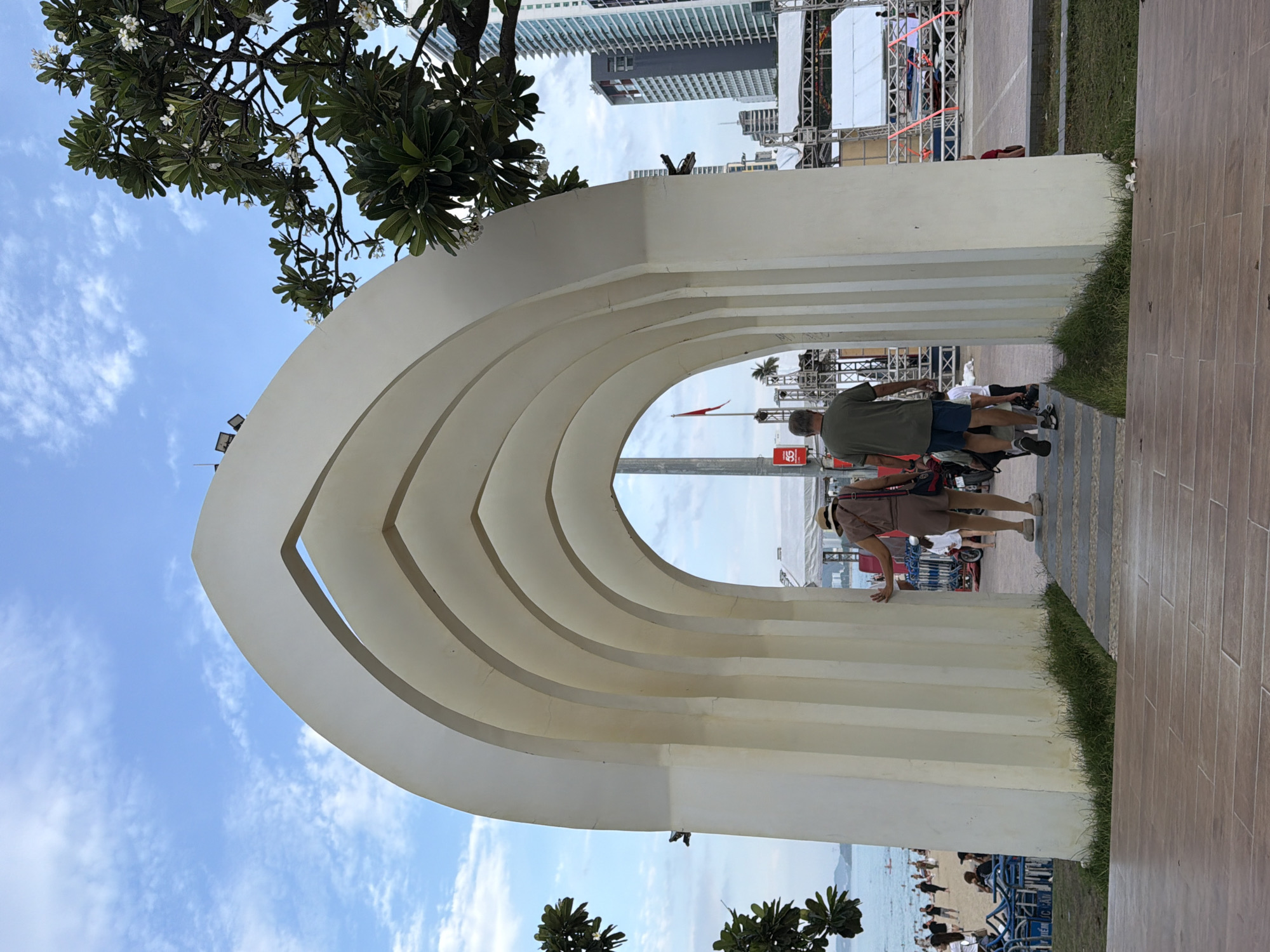 The image shows a wide, cream-colored archway with multiple ribbed arches. Two people are walking through the archway towards the beach. The person in front is a woman wearing a straw hat and a light brown jumpsuit, with a purse slung over her shoulder. Behind her, a man in a green t-shirt and blue shorts walks away from the camera. Beyond the archway, a beach is visible with people, lounge chairs, and the ocean. In the background, there are tall buildings, trees, and a stage structure. The sky is blue with scattered white clouds. This scene is located on the beach promenade in Nha Trang, Vietnam, likely near a public event or performance area given the stage setup.