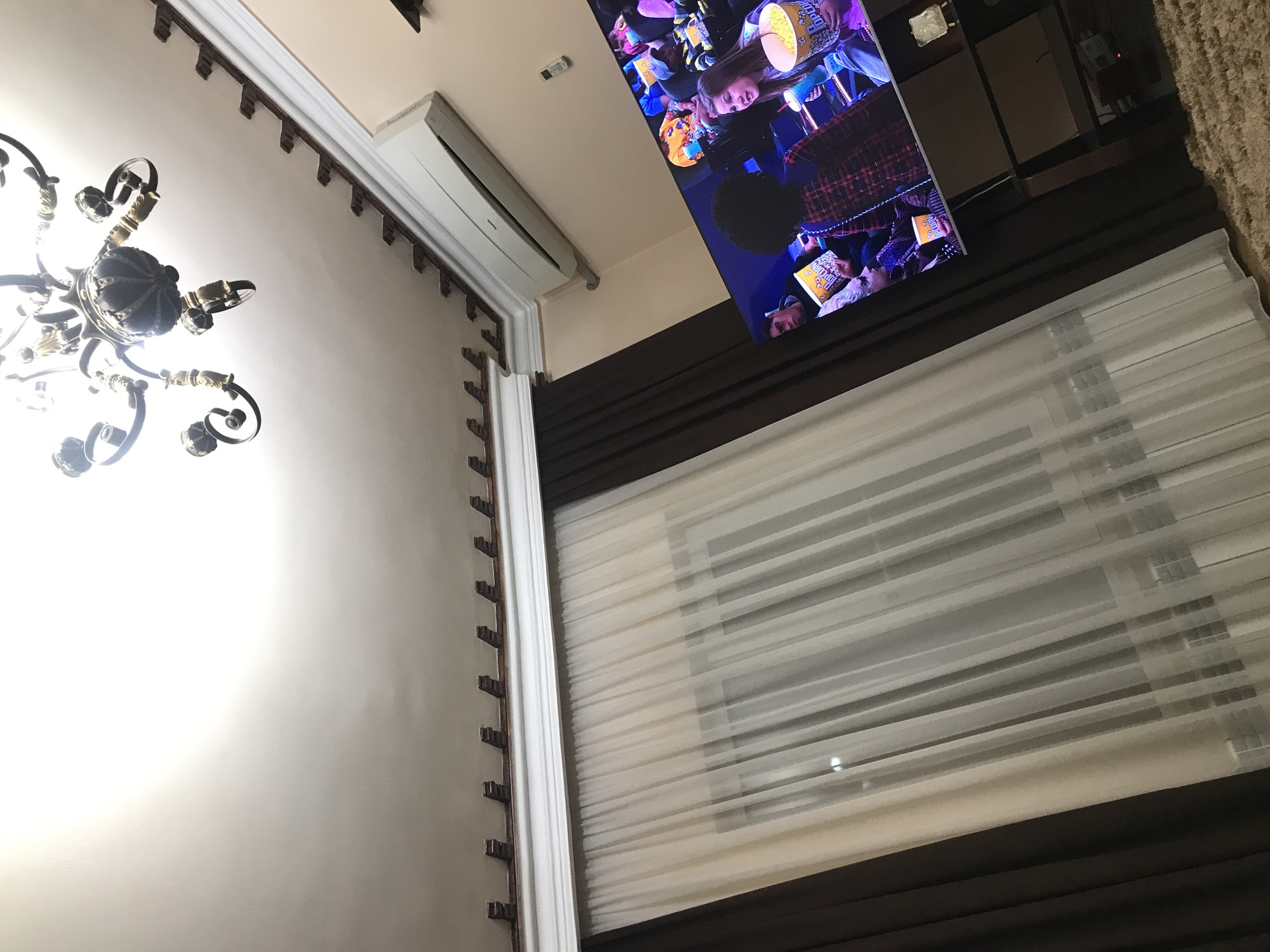 A low-angle shot shows a room with a light beige wall on the left and a large window covered with sheer horizontal blinds on the right. A complex, dark-colored chandelier hangs near the top-left of the frame. A white air conditioning unit is mounted near the ceiling above the window. A flat-screen television is mounted on the wall, displaying a scene from what appears to be a movie theater, with people shown eating popcorn. The window has dark brown curtains above and below the blinds. The image likely depicts an interior space in a residential area. There is no information about the location in Yerevan, Armenia.
