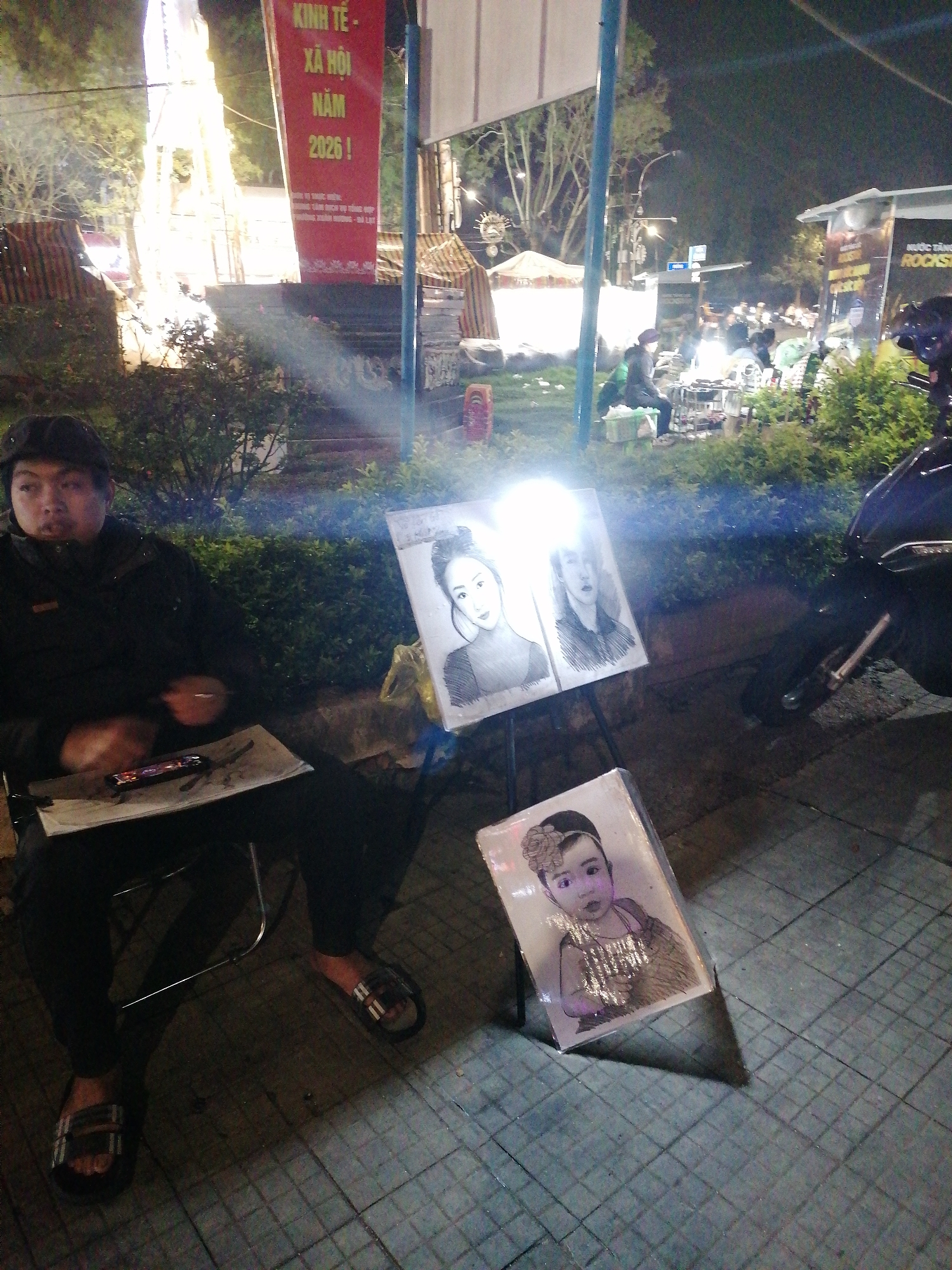 A man is sitting in a metal folding chair on a tiled sidewalk at night in Da Lat, Vietnam. He is wearing a black jacket and black pants, and his feet are clad in sandals. He is holding a small object in his hand and has a mobile phone and a sheet of paper on a board resting on his lap. Two framed drawings of women are positioned on a metal stand to his right, illuminated by a bright light. A third framed drawing of a child is leaning against the stand on the ground. A motorcycle is parked to the right of the drawings. In the background, there is a bustling area with several vendors and people illuminated by bright lights. A tall decorative structure with lights is visible on the left, along with a red banner displaying text in Vietnamese. Shrubs and trees are visible along the sidewalk and in the background.