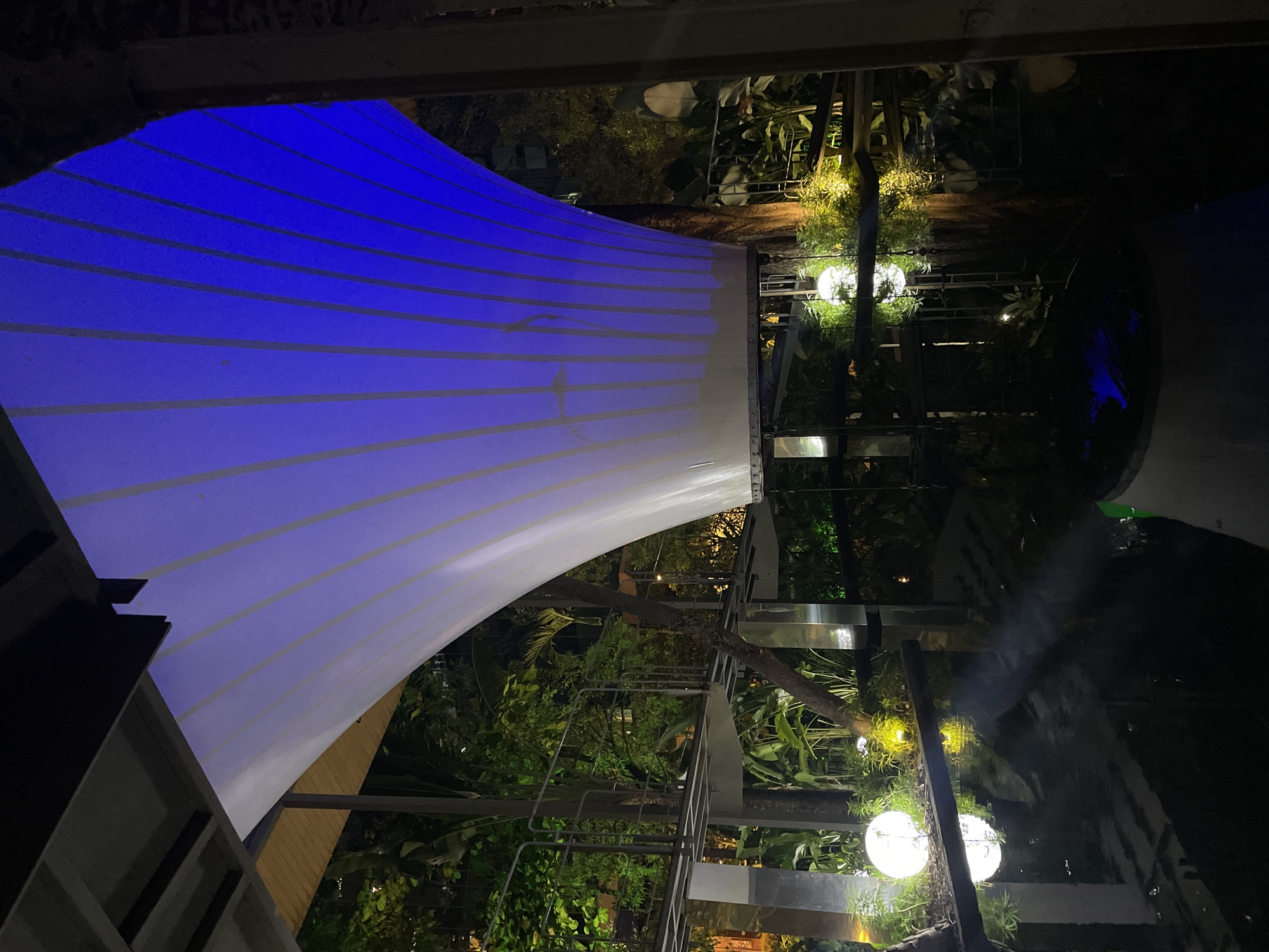 The image shows a nighttime view of an outdoor area in South Jakarta, Indonesia. Overhead is a large white canopy structure with blue lighting on one side, casting a gradient effect. Below the canopy, there is a constructed garden area with various plants and decorative lighting, including spherical lamps. Metal framework and beams are visible, suggesting a modern architectural design. The surrounding area is obscured in darkness, making it difficult to discern further details.