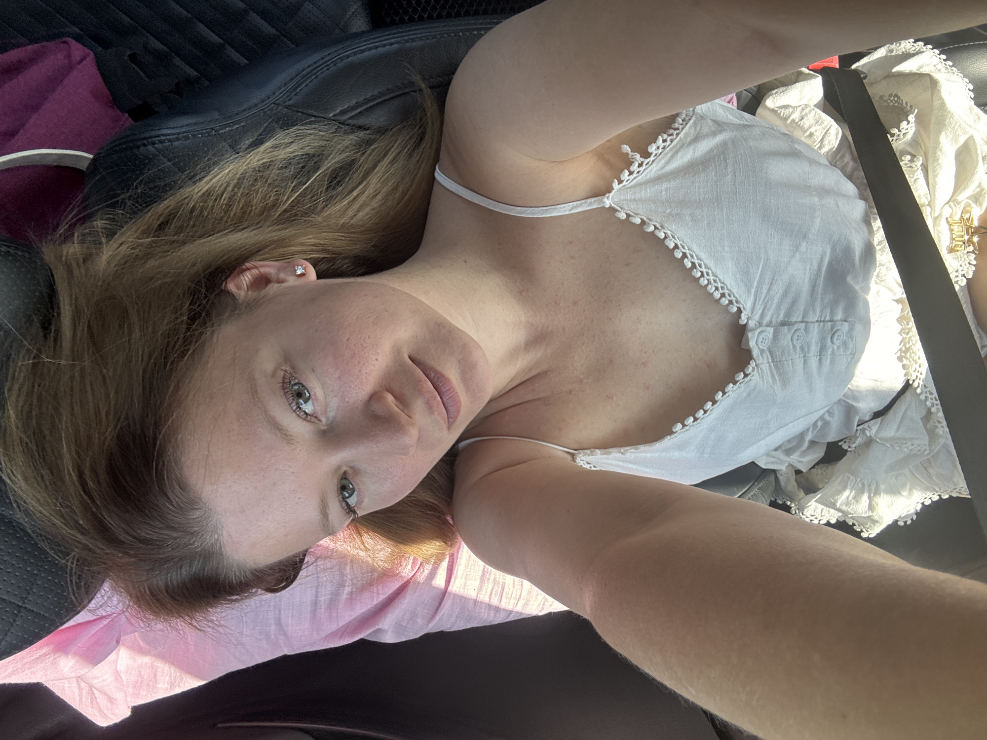 This image is a selfie taken by a woman who appears to be in a car. She is lying down and looking at the camera. She has blonde hair and green eyes. She is wearing a white camisole with lace trim. There is a pink blanket on her left and a black car seat on her right. The image is well-lit and appears to have been taken during the day. The location is not identifiable within the city of Van'ki, Russia.
