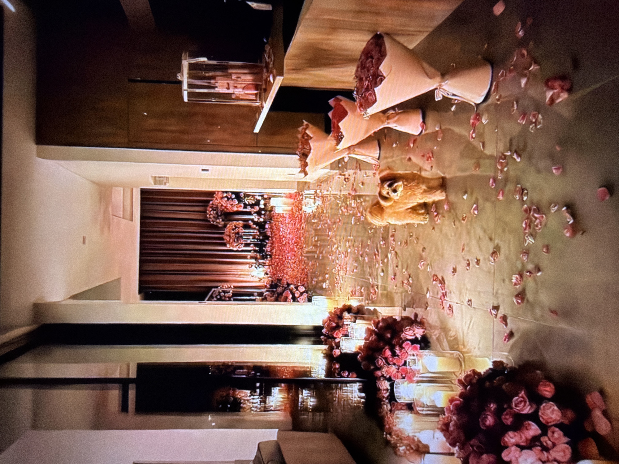 The media file depicts a romantic surprise setup in what appears to be a hallway or entryway of a residence. The scene is lavishly decorated with numerous pink rose petals scattered across the floor, and several bouquets of roses are prominently displayed. Tall, slender glass candle holders with lit candles are strategically placed amongst the flowers, creating a warm and intimate ambiance. In the center of the hallway, a stuffed animal, resembling a dog, is lying down amidst the floral decorations. The lighting suggests it might be evening or nighttime, enhancing the romantic atmosphere. The overall impression is one of a special occasion, likely a romantic proposal or anniversary celebration.