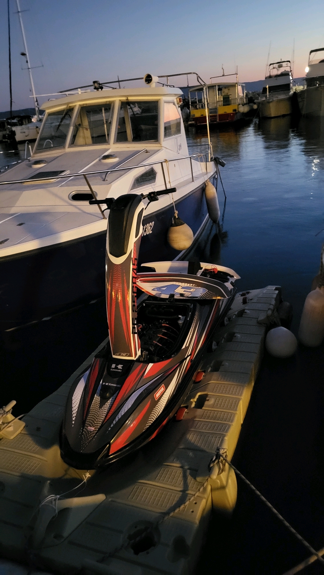 The image depicts a jet ski on a floating dock at dusk in the marina of Općina Tučepi, Croatia. The jet ski, predominantly black with red and white accents and the "Motul" logo visible, is positioned on the right side of the frame. Behind it, a white and blue boat with the number "1362" on its hull is moored. Several other boats are visible in the background, silhouetted against the twilight sky. The water is calm, reflecting the dim light. The scene is serene, with no people visible.