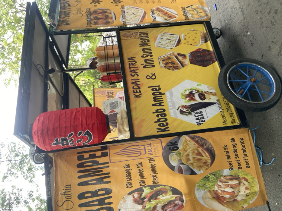 The image shows a food cart in Kecamatan Sidoarjo, Indonesia, located at latitude: -7.441, longitude: 112.705. The cart has a black metal frame and a yellow canvas sign that reads "KEDAI SATRIA Kebab Ampel & Dim Sum Mental". There are several other signs, including one that reads "Satra Bab Ampel". The cart is selling a variety of food items, including kebabs, fries, and dim sum. There are several pictures of food on the signs, as well as a price list for the different items. There is also a red paper lantern hanging from the top of the cart. The cart is parked on the side of the road, and there are trees and other foliage in the background. A person is visible in the background wearing a blue shirt, seemingly manning the cart.
