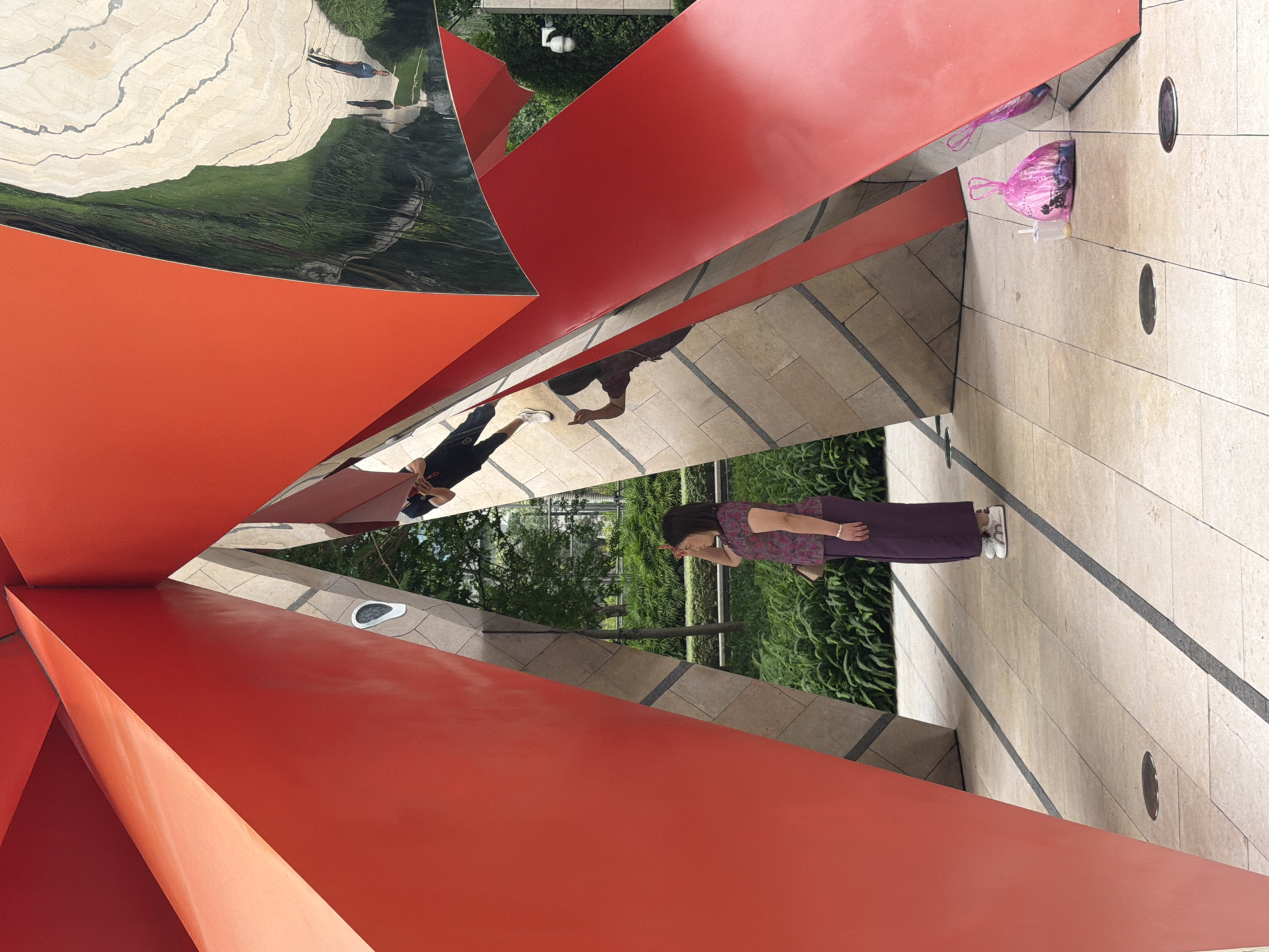 A woman in a purple patterned top and long purple skirt stands in a courtyard, shielding her eyes with her right hand. She wears white sneakers and has a small purse on her right hip. The courtyard is paved with light-colored square tiles with dark gray grout lines. The scene is framed by large, angular, bright red abstract sculptures. Some of these sculptures have reflective surfaces that show distorted images of the surroundings, including people and greenery. To the right of the woman, a pink plastic bag and a plastic cup with a straw are on the ground. In the background, lush green bushes and trees are visible. The overall impression is one of a modern urban art installation.