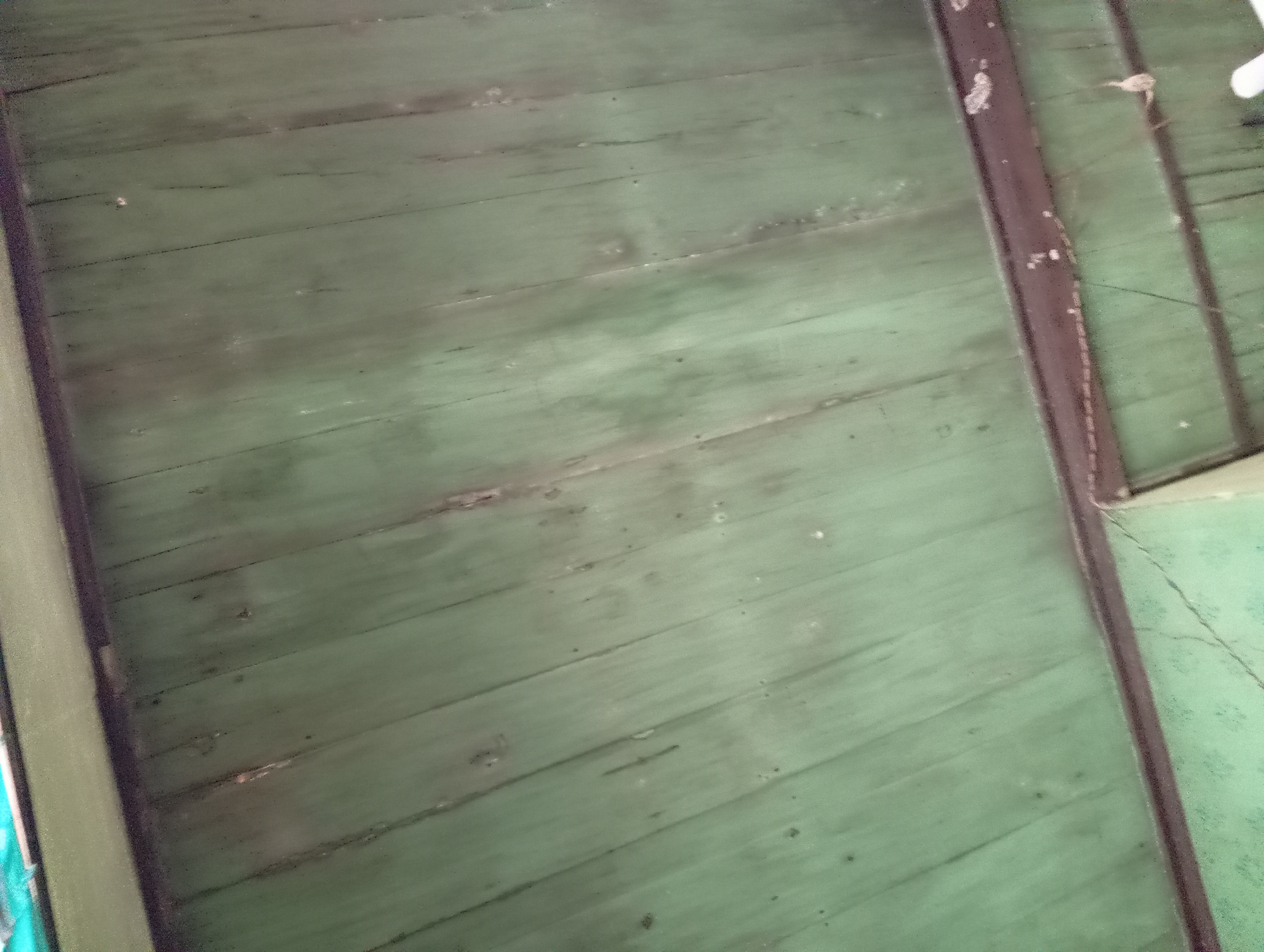 The image shows a close-up view of a weathered, green wooden ceiling, possibly part of a building in Rejosari, Indonesia. The planks are oriented diagonally from the top left to the bottom right of the frame. The wood shows signs of age, with visible grain, cracks, and some darker spots. A dark brown metal beam runs horizontally across the lower portion of the image, separating the ceiling from what appears to be a wall or another section of the structure below, which is also green. There are faint spiderwebs visible on the beam and the lower green surface. The lighting suggests an indoor setting with ambient light.