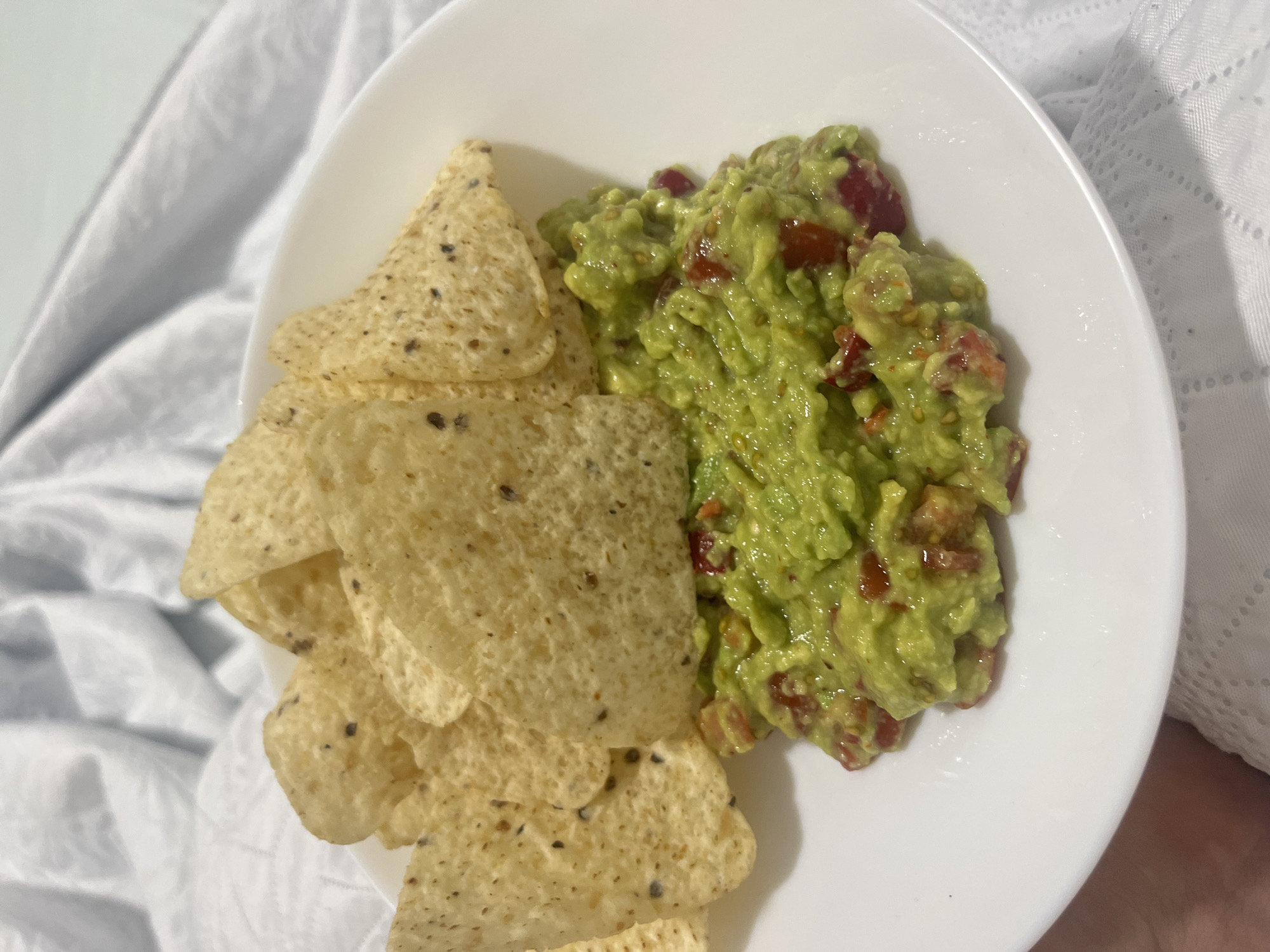A white plate containing a pile of tortilla chips and a serving of guacamole sits on what appears to be a white bedsheet. The guacamole, predominantly green, contains visible diced red tomatoes. The chips are light tan in color and speckled with darker flecks. No people are visible in the frame. The setting appears to be indoors. There is no information that it is in the city of Mogán, Spain.