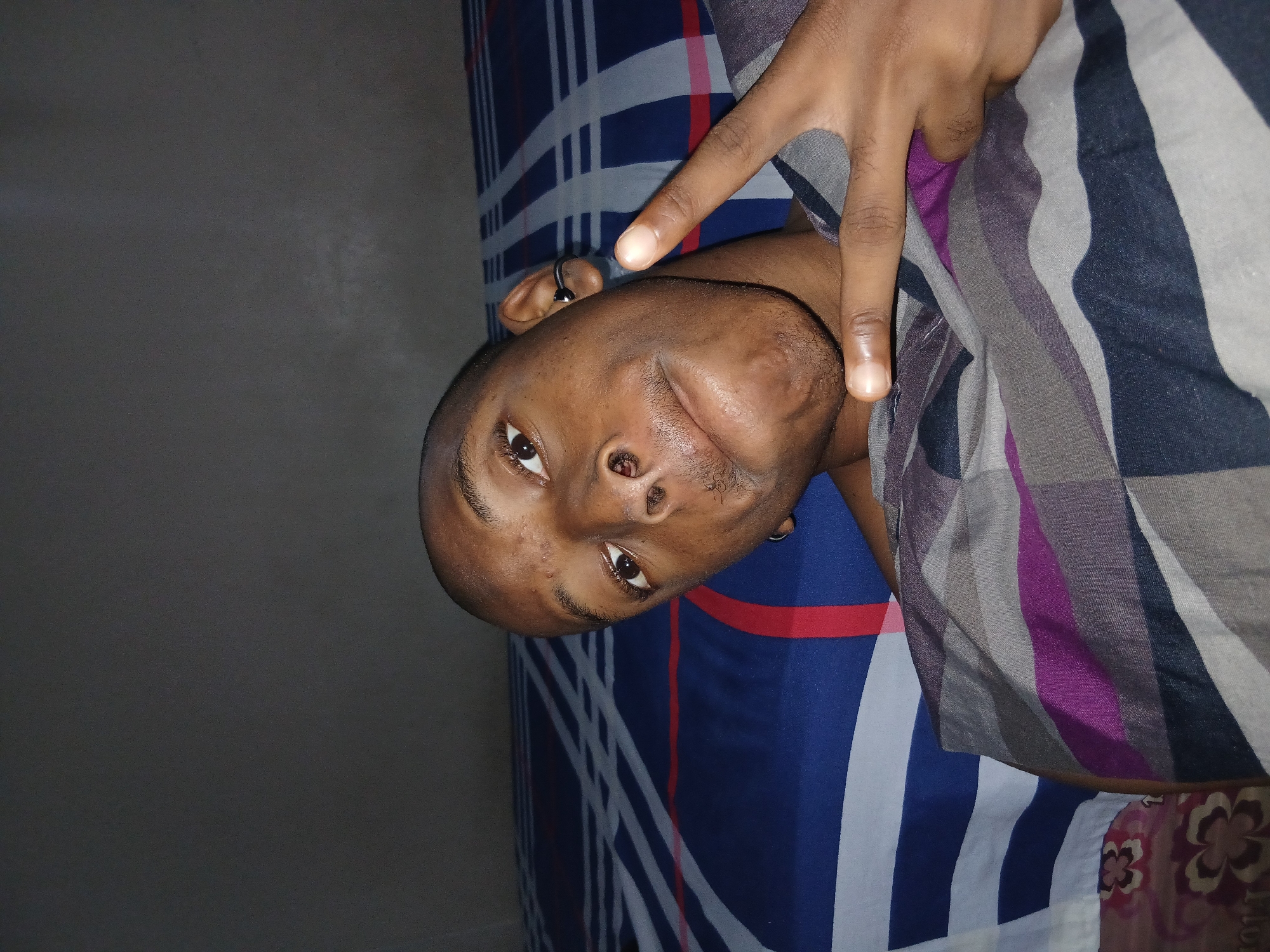 A close-up selfie shows a bald Black person lying on their back in bed, with their head tilted slightly. They are looking at the camera with their right eye more visible than their left. A peace sign is being made with their right hand, fingers extended upwards, obscuring part of their face. The person is wearing a grey, purple, and black striped shirt. They have a small hoop earring in their left ear. The background consists of a dark wall and a bed with striped blue, white, and red bedding. The scene appears to be indoors, likely a bedroom. No specific location in Lagos, Nigeria can be identified from the provided image.