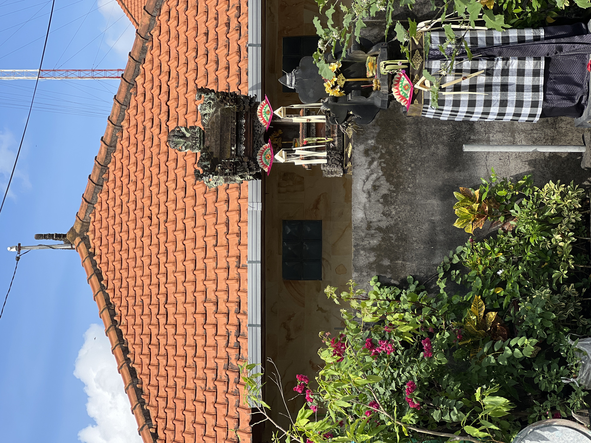 In the city of North Kuta, Indonesia, this image captures a Balinese shrine adorned with offerings. The shrine, made of dark stone, is wrapped in a black and white checkered cloth. It is decorated with colorful fans, flowers, and incense. Behind the shrine, a building with a terracotta tile roof is visible, with lush green foliage in the foreground. The sky is a bright blue with scattered clouds.