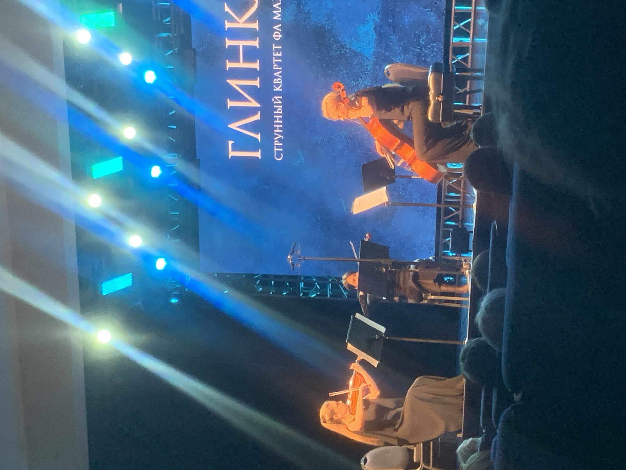 The image shows a string quartet performing on a stage in Izhevsk, Russia. The backdrop displays the text "ГЛИНКА" and "СТРУННЫЙ КВАРТЕТ ФА МАЖ," likely indicating the performance of a work by the composer Glinka. The quartet consists of a cellist, two violinists, and a violist. They are seated and focused on their sheet music, performing on their instruments. There are stage lights with blue and white beams illuminating the scene. The audience is partially visible in the foreground, sitting in rows of seats. The image appears to be taken from an audience perspective.