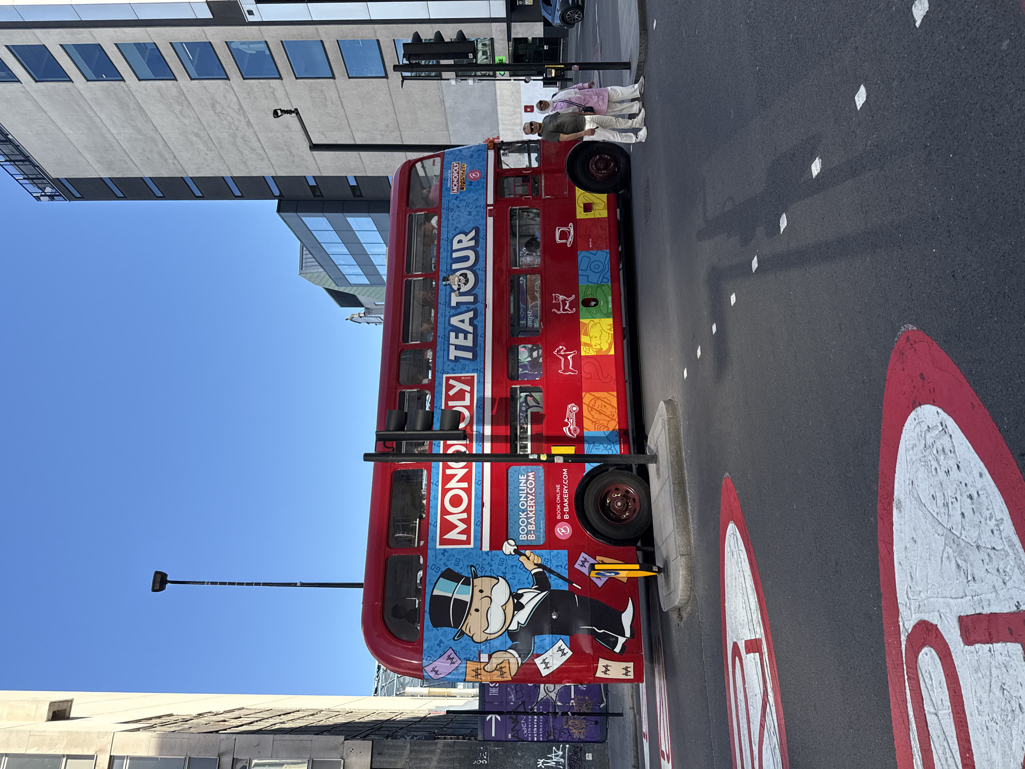 Here is a description of the image:
The image shows a red double-decker bus parked on a street in London, United Kingdom. The bus is branded with "MONOPOLY TEA TOUR" in large letters and features the Monopoly mascot on the side. There are people standing near the bus, suggesting that it might be stopped or operating as a tour bus. The bus is located in an urban environment with buildings on both sides. The sky is clear and blue. Road markings are visible on the street, including speed limit signs. The image is rotated 90 degrees counterclockwise.
