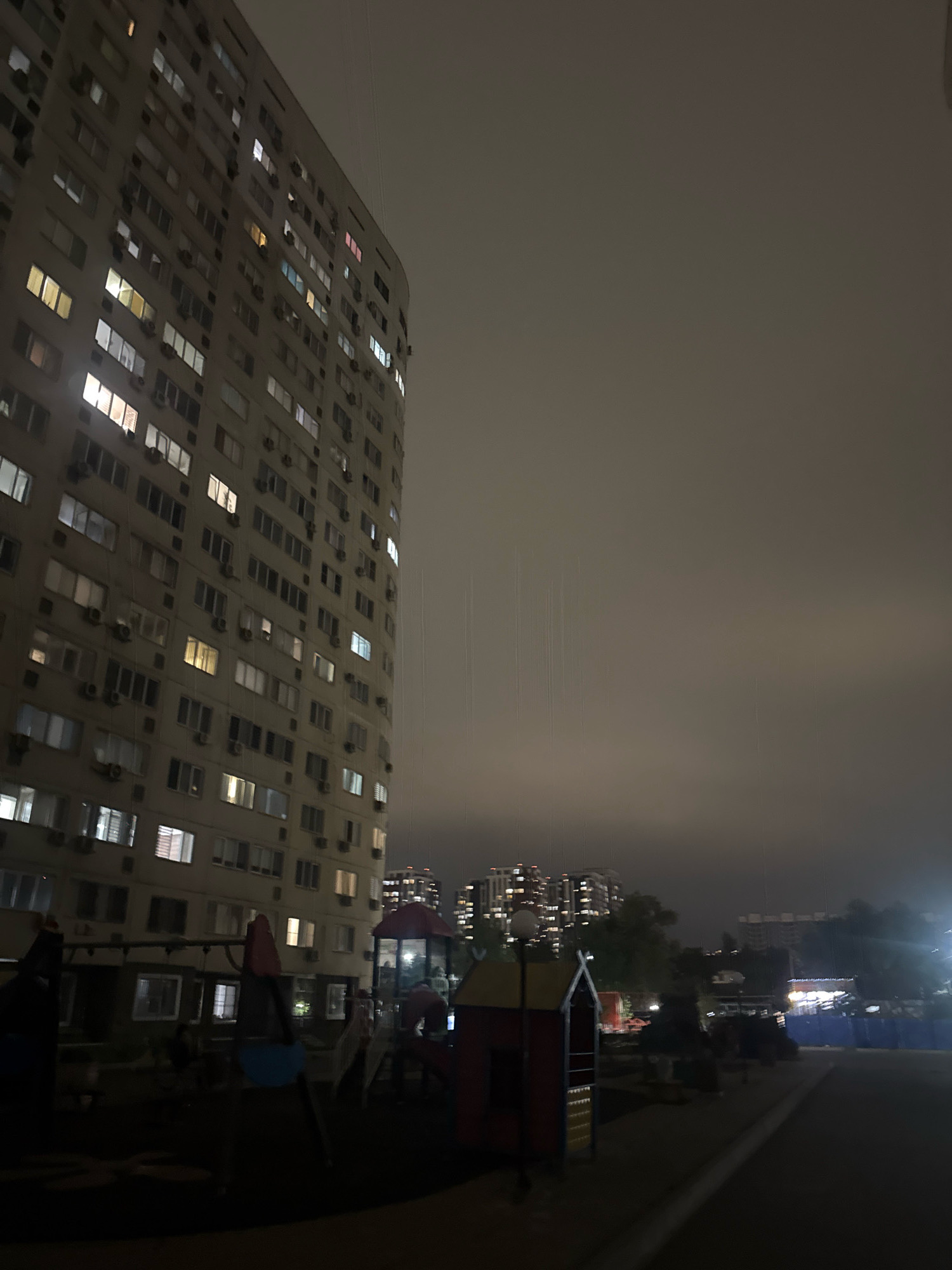 This is a night-time photograph taken in a residential area of Almaty, Kazakhstan. The primary subject is a tall, multi-story apartment building on the left side of the frame. Many windows of the building are illuminated, indicating that people are inside. In the foreground, there is a children's playground with various structures, including slides and a small house-like play area. In the background, other apartment buildings and trees are visible, partially obscured by darkness and atmospheric haze. The sky is overcast and dark, suggesting it might be raining, as indicated by faint vertical lines visible in the upper right portion of the image. The overall scene depicts a typical urban residential environment after dark.