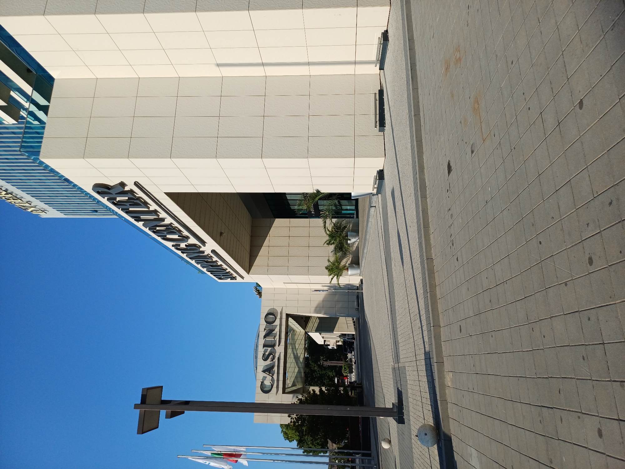 The image shows the entrance to a shopping center and casino in Varna, Bulgaria. The architecture is modern, with beige tiled walls and large lettering spelling out "SHOPPING CENTER" and "CASINO". There are flags flying to the left of the "CASINO" entrance. A lamppost stands prominently in the foreground. The ground is paved with grey tiles. There are potted plants visible inside the entrance. The sky is a clear blue.