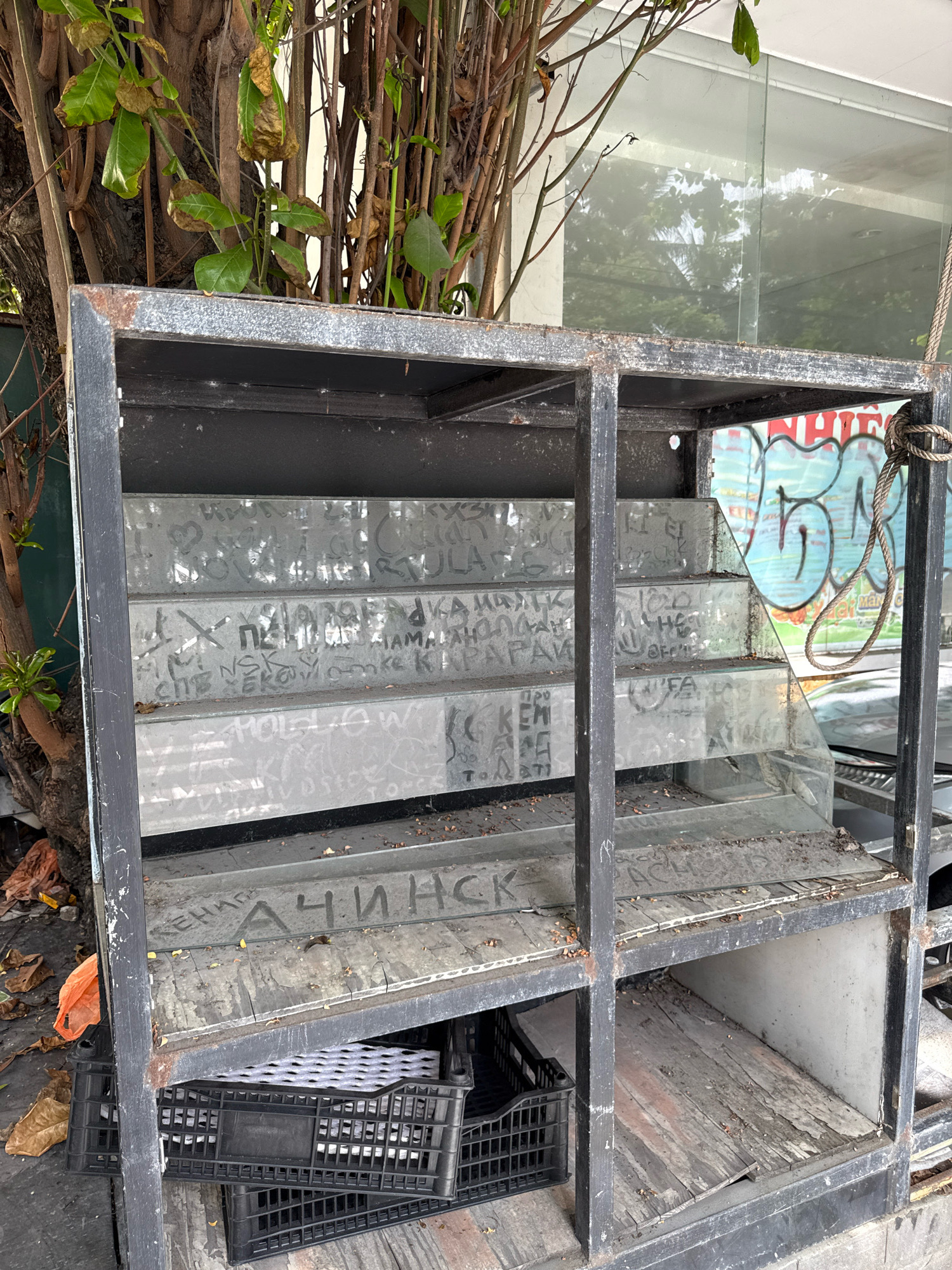 This image depicts an outdoor display shelf in Nha Trang, Vietnam, likely outside a shop or stall. The shelf unit is constructed of dark metal, showing signs of rust and wear. It has multiple tiered shelves, some of which are made of glass and others of wood. The glass shelves are covered in graffiti and scribbled writing. On the bottom shelf, two black plastic crates are stacked, one containing white material that appears to be packaging or padding. To the left of the shelf, there are fallen leaves and an orange plastic bag. Behind the shelf, a tree with branches and green and brown leaves provides some shade, and a glass-fronted building with reflections of trees and sky is visible. The overall scene suggests an abandoned or disused commercial space, exposed to the elements and subject to graffiti.