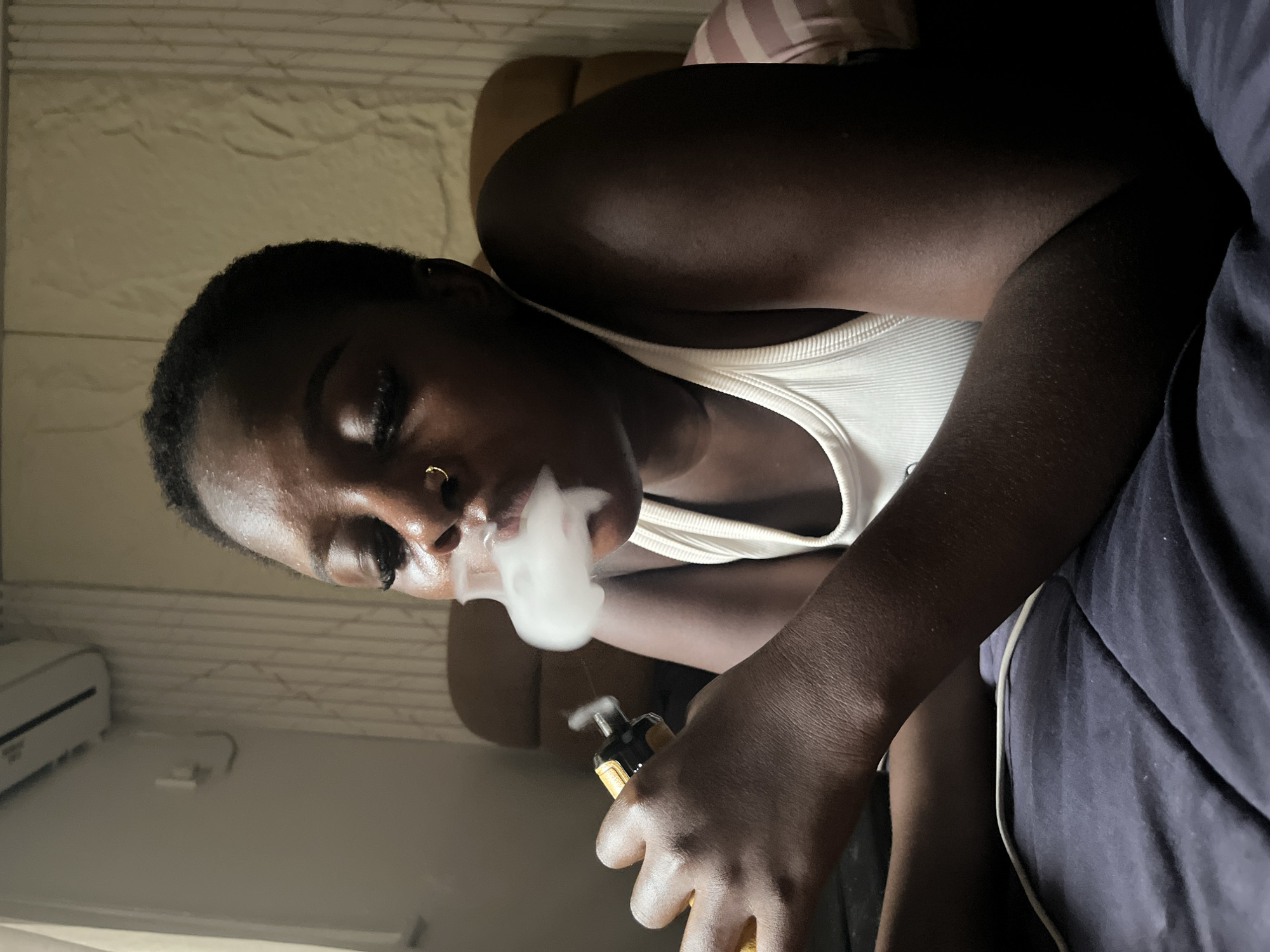 A person with dark skin and a short haircut is lying down on a dark blue surface, possibly a bed or couch. They are wearing a white tank top and have a gold hoop nose ring. In their right hand, they are holding a yellow and black electronic device, from which a plume of white vapor is being exhaled from their mouth. The person's eyes are closed. In the background, there is a textured wall with vertical lines, and what appears to be an air conditioner unit mounted high on the wall to the left. The lighting in the scene is dim.