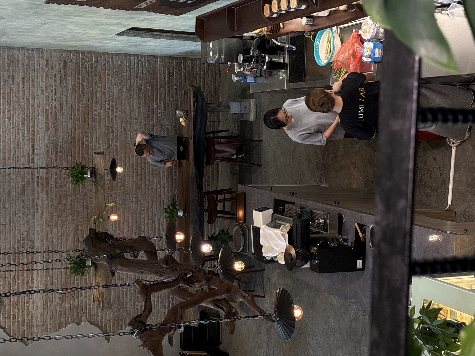 This is an overhead shot of a modern cafe in Da Nang, Vietnam, with exposed brick walls and industrial-style lighting. In the background, a man with short hair stands at a long wooden table, looking at his phone. In the foreground, two women work behind a counter. One woman, wearing glasses and a grey t-shirt, appears to be preparing something. The other woman, wearing a black t-shirt with "LUMI LAB" printed on the back, is also working at the counter, which is equipped with a coffee machine, grinder, and various cleaning supplies. The cafe's interior design features large, decorative chains hanging from the ceiling and a prominent wooden structure that adds an organic element to the space. The overall atmosphere suggests a bustling urban cafe setting.