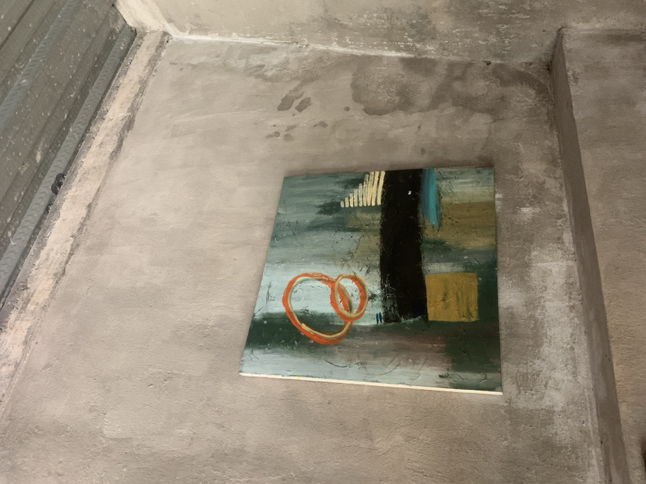 The image shows a corner of an interior space with a rough, textured concrete wall. A painting is hung on the wall. The painting depicts abstract geometric shapes and colors, including orange, green, black, yellow, and blue. The painting is mounted on a white canvas. The corner is located on the ground floor of a building in the village of Seyegan, Kecamatan Piyungan, in the Special Region of Yogyakarta, Indonesia. The latitude and longitude coordinates are -7.825, 110.453.