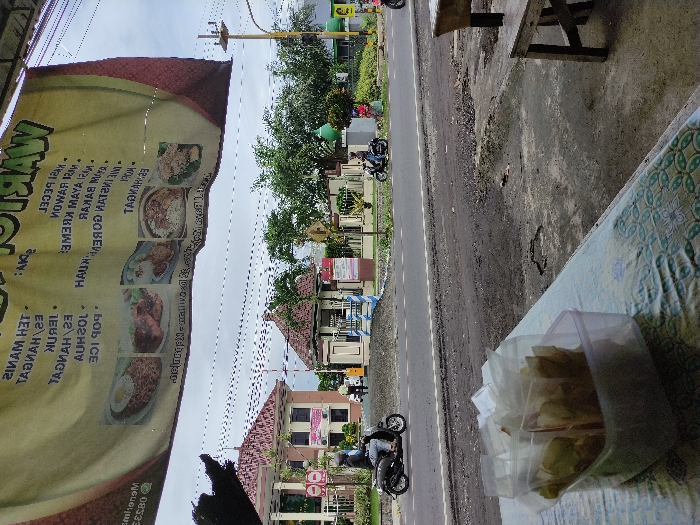 This image captures a vibrant street scene in Kepanjen, Indonesia, viewed from what appears to be a roadside food stall or warung. The scene is bright, suggesting daytime with an overcast sky.

In the immediate foreground, a large yellow and brown banner, likely for a food establishment, dominates the left side. The banner, oriented vertically in the original image, displays the name "WARUNG TOGOR" in large green lettering. Below the name, a menu lists various Indonesian food and drink items, accompanied by images of dishes like fried rice with an egg, fried chicken, and noodle soup. Visible menu items include "TEH MANIS," "ES JERUK," "POP ICE," "NASI PECEL," "MIE AYAM," "NASI BAKAR," "RAWON MANGKOK" (or "MAWANG"), "NASI GORENG KUAH," and "MIE AYAM KREMES." A phone number starting with "0852..." is also partially visible at the bottom of the banner. To the right, a table draped with a light blue and white patterned tablecloth holds a clear plastic container filled with individually wrapped snacks, possibly crackers or chips.

The midground features a paved road with a distinct white dividing line, where several motorcycles are visible, some parked and others in motion, indicating active traffic. Beyond the road, the background reveals lush green trees, a grassy area that might be a park, and several buildings. One building stands out with a red-tiled roof, while another, farther back, is a multi-story structure with various signs. The overall setting suggests a bustling semi-urban or suburban area. No specific human emotions are discernible, as the people are mostly distant or obscured.