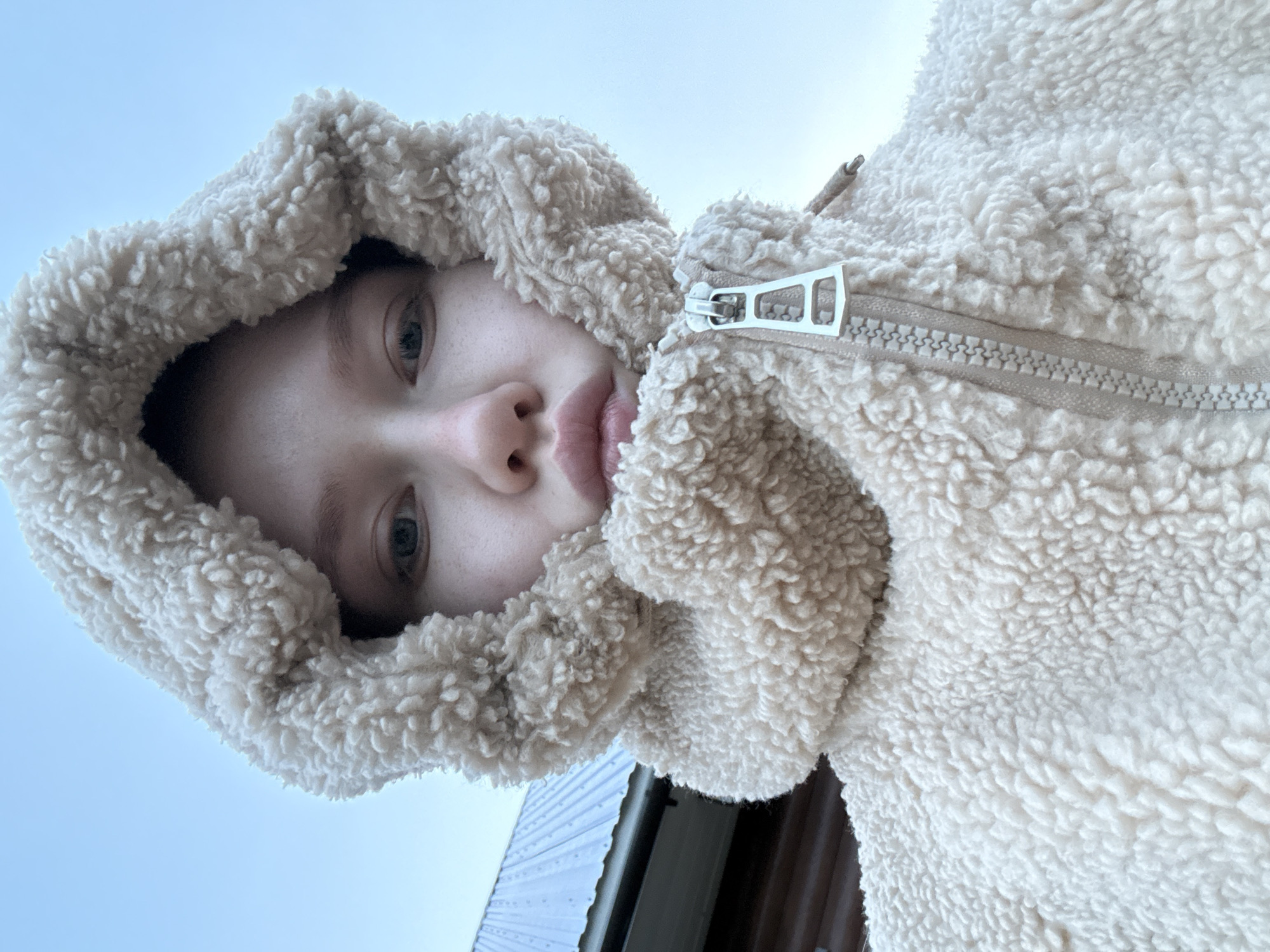 A young person is centered in the frame, looking upwards at the camera from a low-angle perspective. The person is wearing a cream-colored fleece jacket with the hood up. The jacket's zipper is partially open, with the zipper pull visible.
In the background, partially visible, is a building with a corrugated metal roof. The sky is overcast and light blue, filling the majority of the upper frame. There is insufficient information to ascertain the location of the scene as Zhigalki, Russia.