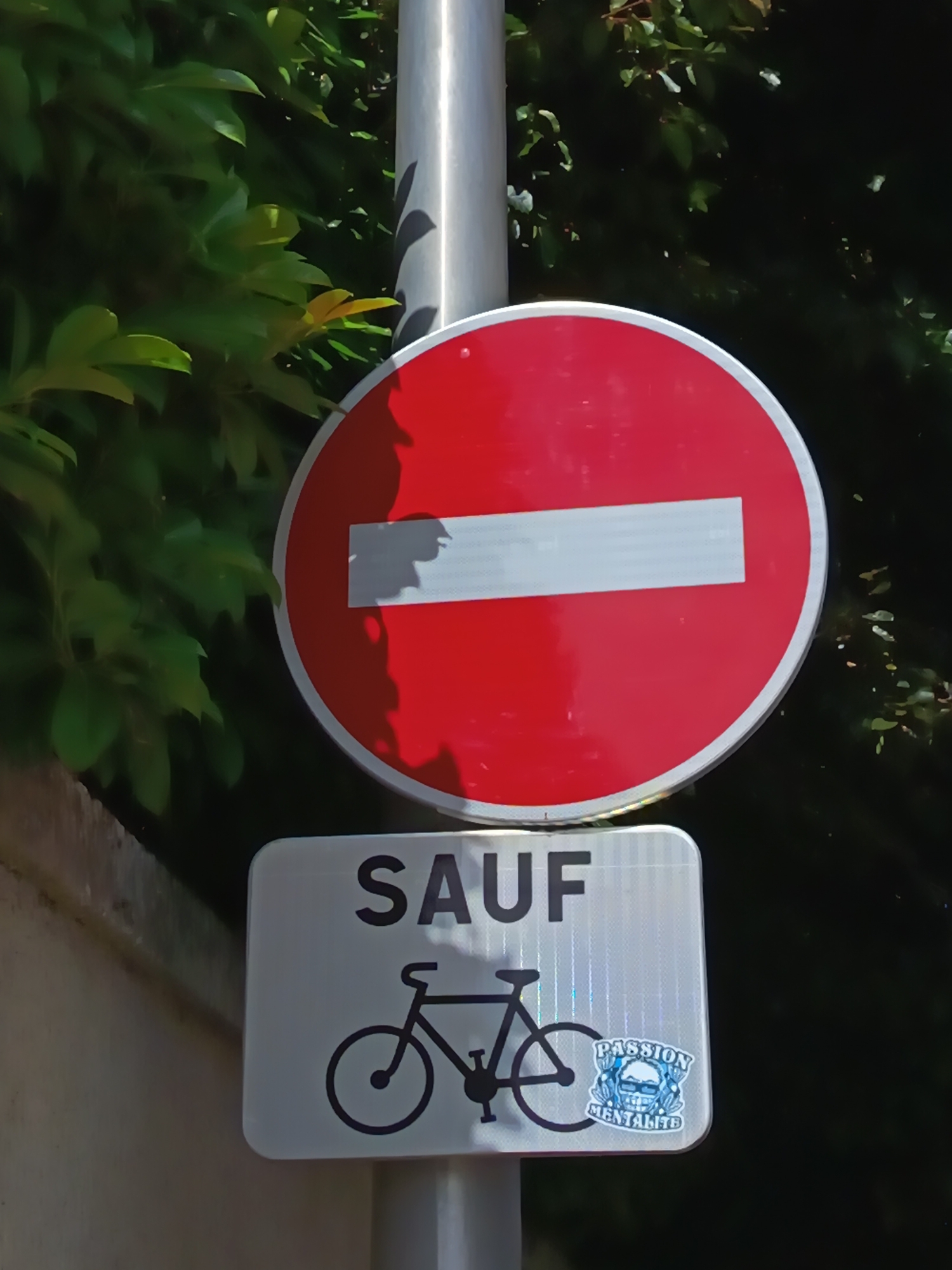 The image shows a close-up view of a traffic sign in Périgueux, France. The sign consists of two parts. The upper part is a circular red sign with a white horizontal bar, indicating "no entry" or "do not enter." Below this is a rectangular white sign with the word "SAUF" (French for "except") in large black letters. Beneath "SAUF" is a black silhouette of a bicycle. To the right of the bicycle, a small sticker is attached to the white sign. This sticker is blue and white and features a skull wearing sunglasses with the words "PASSION MENTALITE" written below it. The signs are mounted on a metal pole. The background consists of lush green foliage, suggesting an outdoor, possibly park-like or tree-lined street setting. The lighting indicates it is daytime, with sunlight casting shadows.