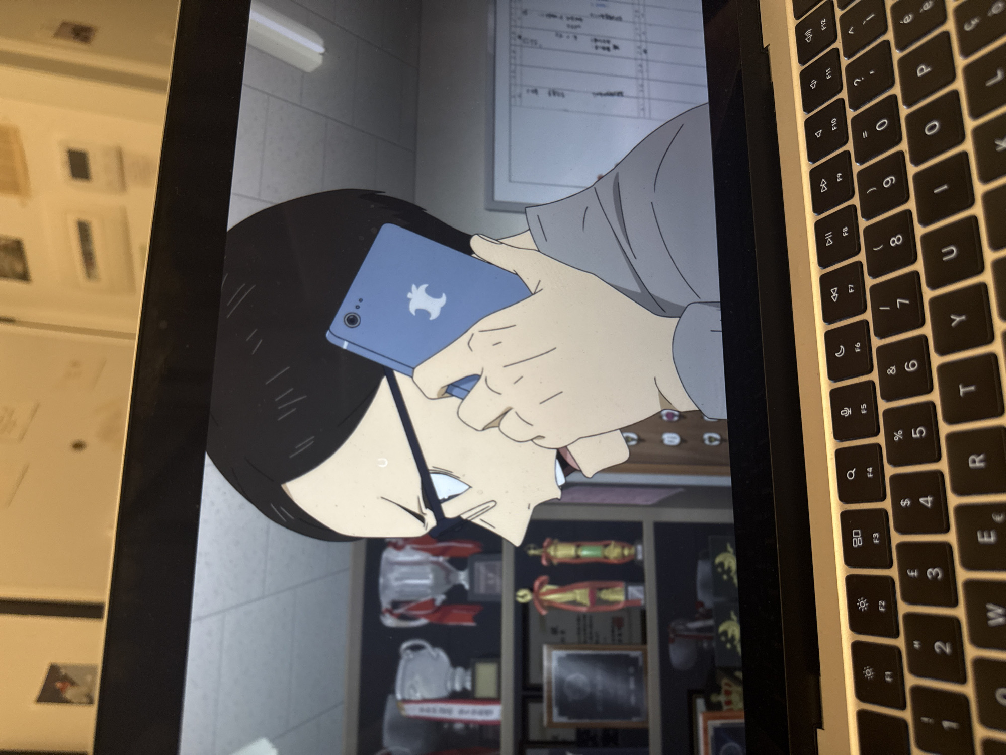 A laptop screen displays an animated scene featuring a male character with black hair and glasses, holding a light blue smartphone with an Apple logo to his ear. The character appears stressed or concentrated, indicated by sweat marks on his temple. The phone's back also shows two camera lenses. In the background of the animated scene, there are shelves filled with trophies and awards, and a whiteboard with illegible text or drawings, suggesting an indoor setting like an office or classroom.
To the right of the screen, part of a laptop keyboard is visible, characterized by silver keys with black characters, typical of an Apple MacBook. Visible keys include the function row (F1-F12) with icons for screen brightness, expose, launchpad, keyboard backlight, and media controls (play/pause, volume up/down, mute). Alphanumeric keys such as 'Q', 'W', 'E', 'R', 'T', 'Y', 'U', 'I', 'O', 'P', and number keys '1' through '0' are also discernible, along with various symbols including '£', '$', '%', '&', '*', '(', ')', '-', '+', '=', and common punctuation. The image itself is rotated, showing the screen and keyboard at a 90-degree angle relative to standard viewing. The location context provided is Milan, Italy.