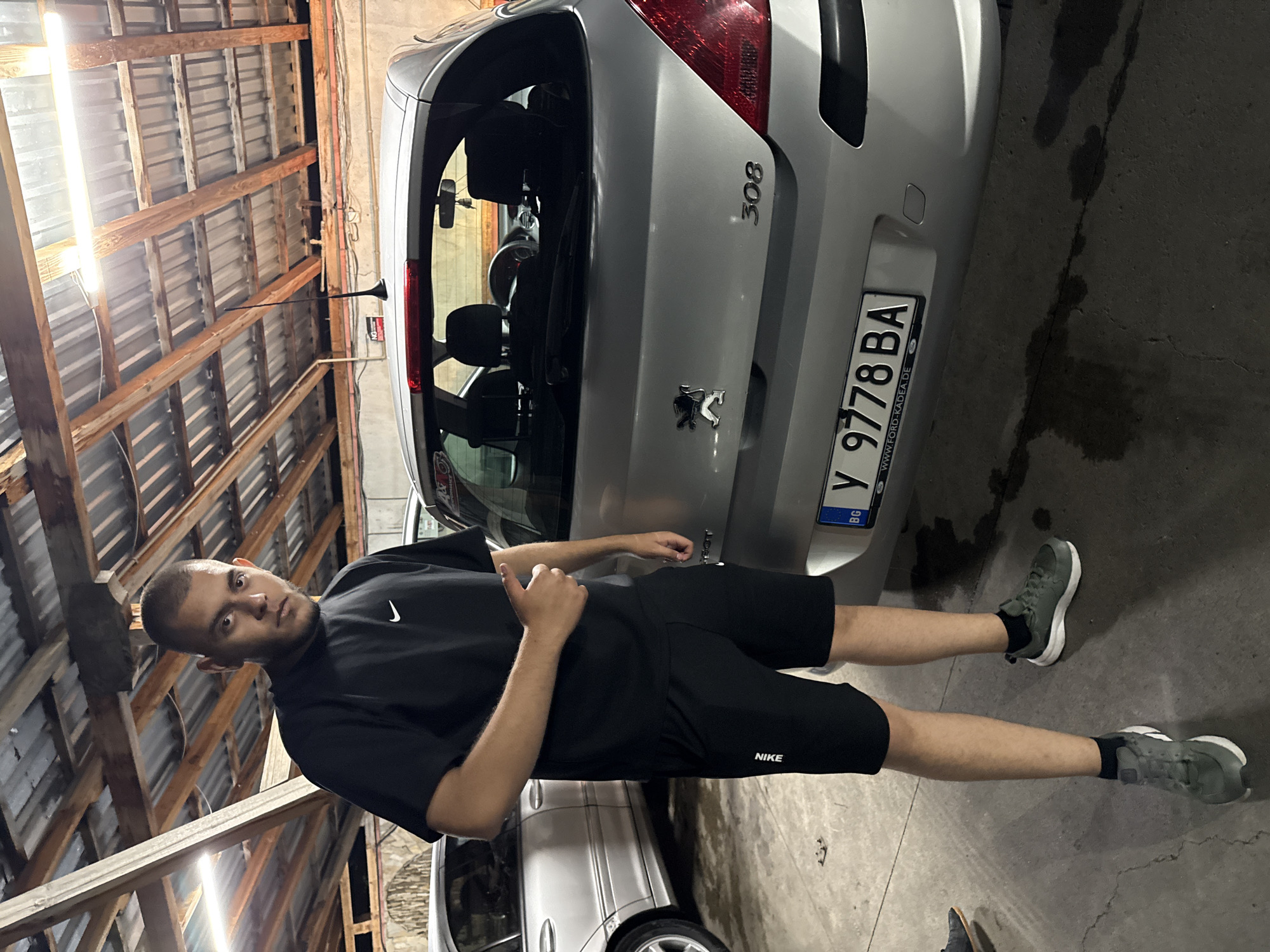 A man stands in a garage next to a silver Peugeot 308. The garage has a corrugated metal ceiling supported by wooden beams. The car's license plate is Y 9778 BA, indicating it is registered in Bulgaria. The man is wearing a black t-shirt with a Nike logo and black Nike shorts. He has short, dark hair. His feet are wearing gray sneakers. The location in the city of Elhovo, Bulgaria is not possible to ascertain from the picture.