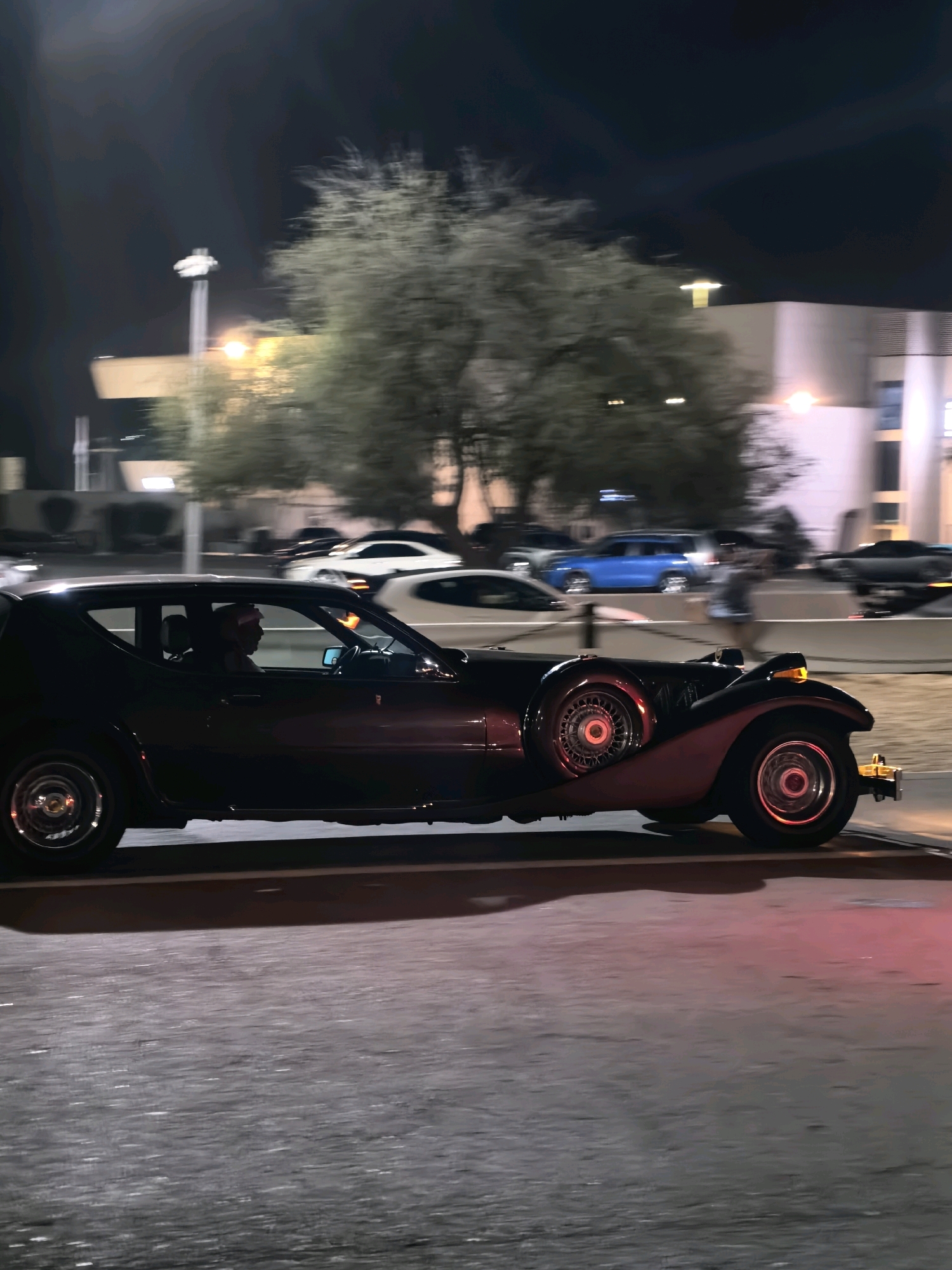 The image depicts a classic black convertible car, possibly a Zimmer Golden Spirit or similar replica, driving on a wet asphalt road at night in Abu Dhabi, United Arab Emirates. The car features a prominent external spare tire on the passenger side, chrome accents on the wheels, and visible headlights and taillights. A person wearing traditional Arabic headwear is visible in the driver's seat. In the background, trees with sparse foliage are illuminated by streetlights, and modern buildings with architectural lighting are present. Other vehicles, including a blue SUV and white sedans, are also visible on the road, suggesting an urban environment. The overall impression is that of a nighttime street scene in a well-lit city.