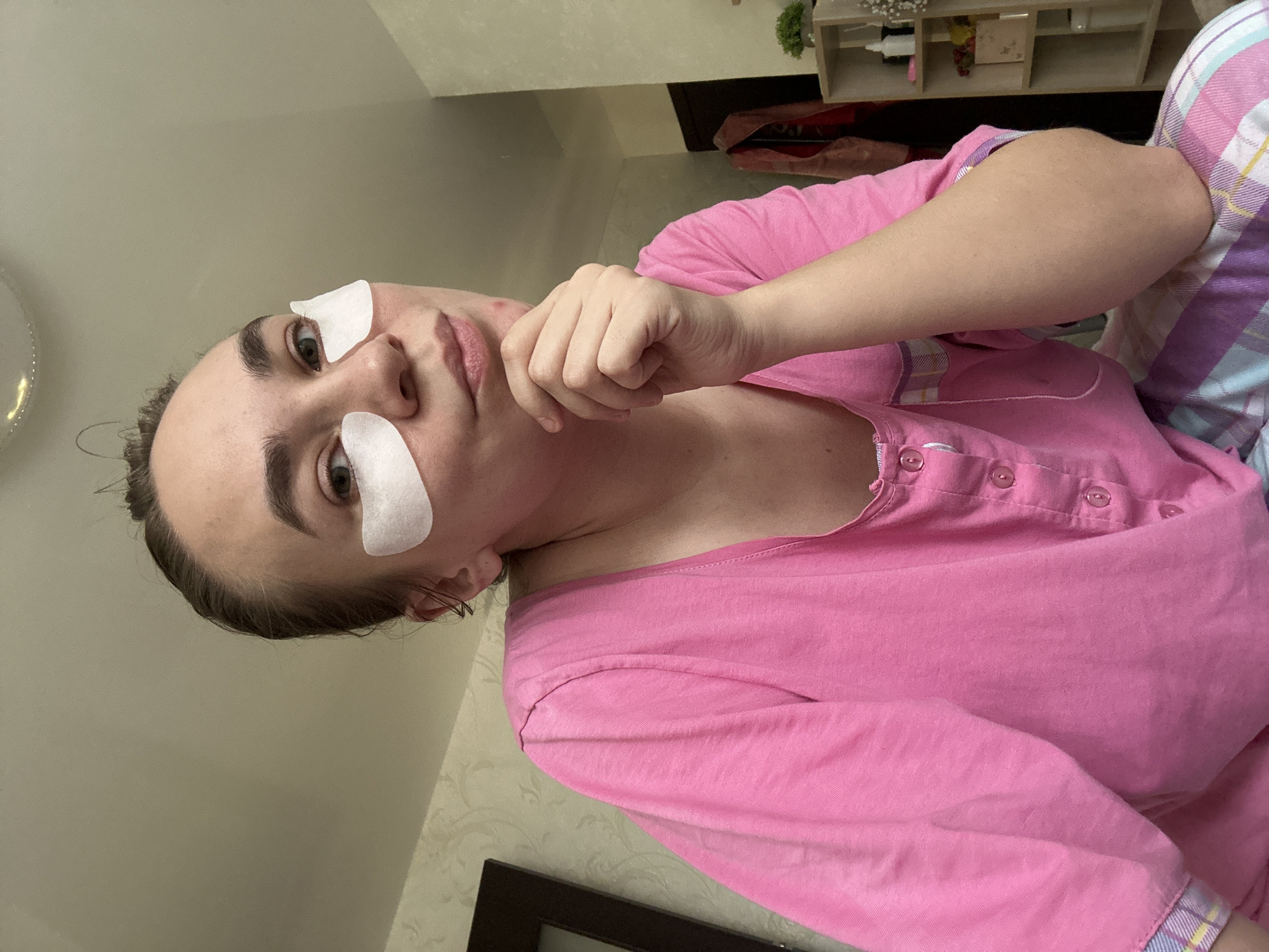 A woman is shown from a low-angle point of view. Two white cosmetic patches are placed under her eyes. She is wearing a pink, button-down shirt, and plaid pajama pants. She is touching her face with her right hand. In the background, there is a wall with a floral wallpaper pattern and some shelves. The location, Uva, Russia, cannot be inferred from the image.